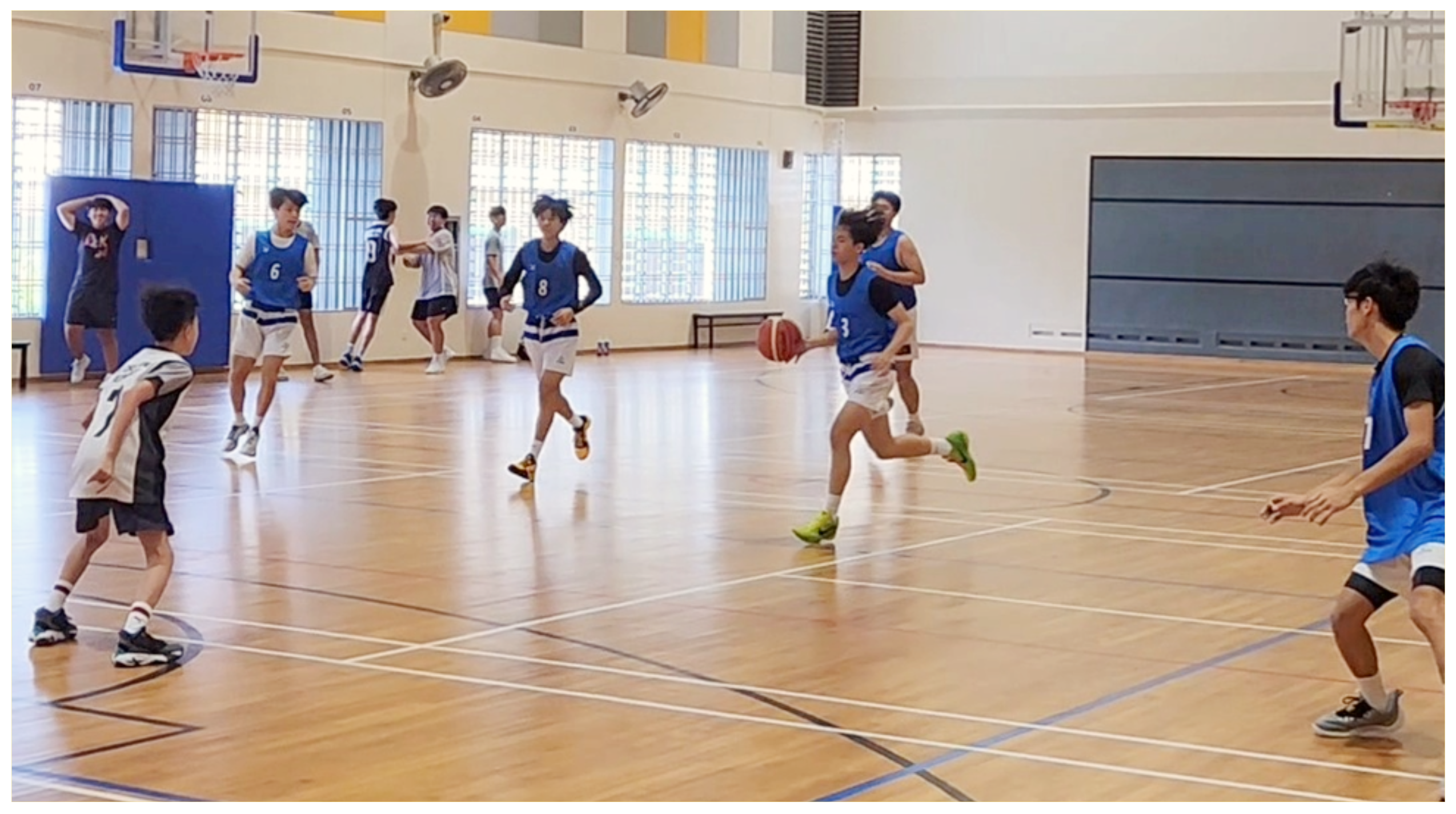 Young players in blue and white jerseys play basketball on an indoor wooden court with hoops visible.