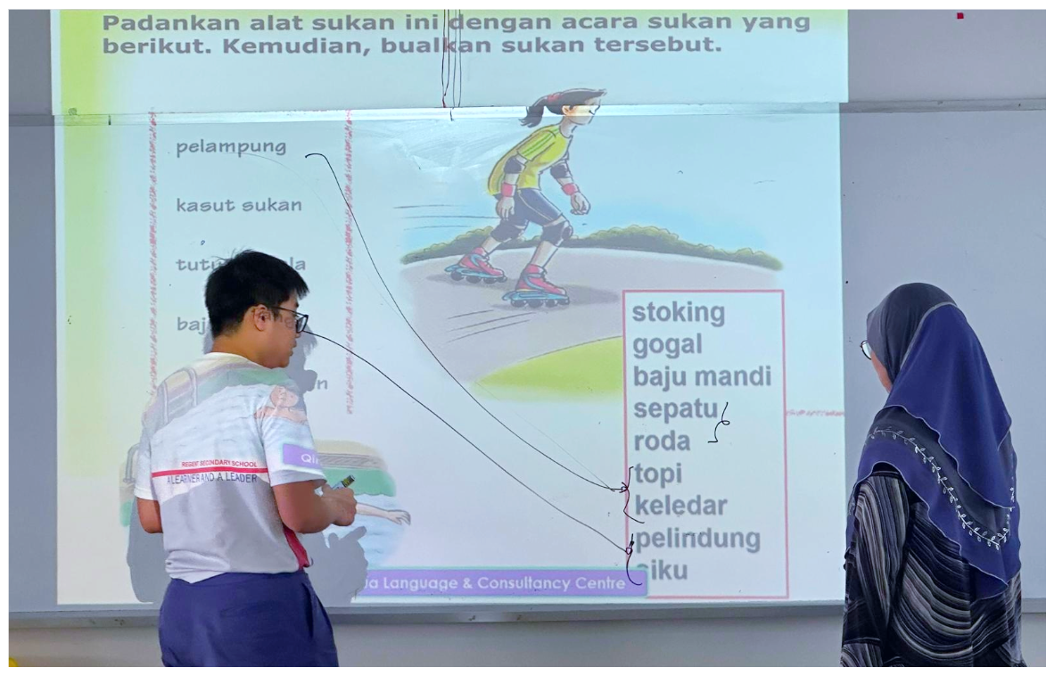 A student and teacher stand before a projected Malay language worksheet showing a rollerskating activity matching exercise.