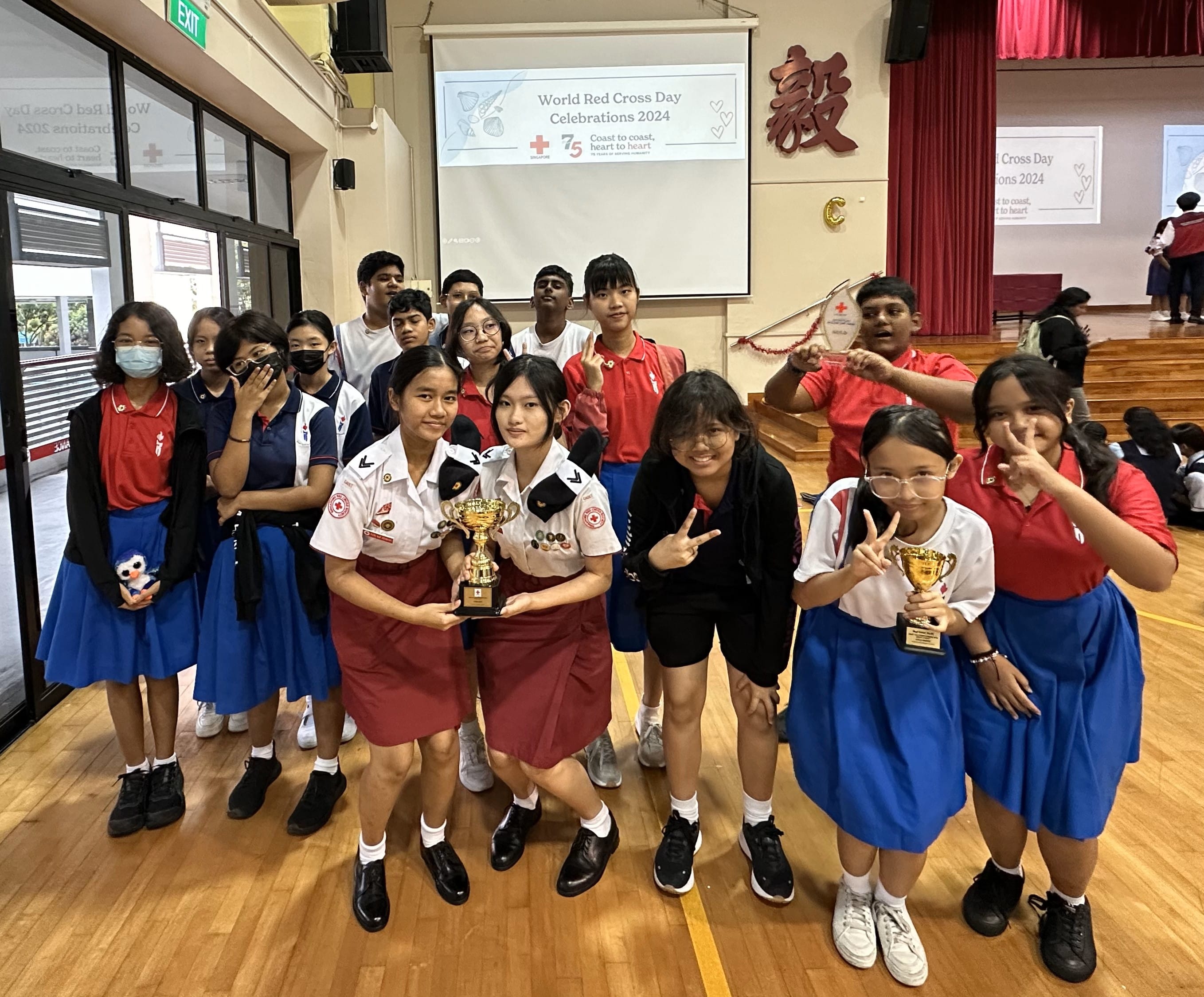 Students in school uniforms hold gold trophies at a World Red Cross Day Celebrations 2024 event in a school hall.