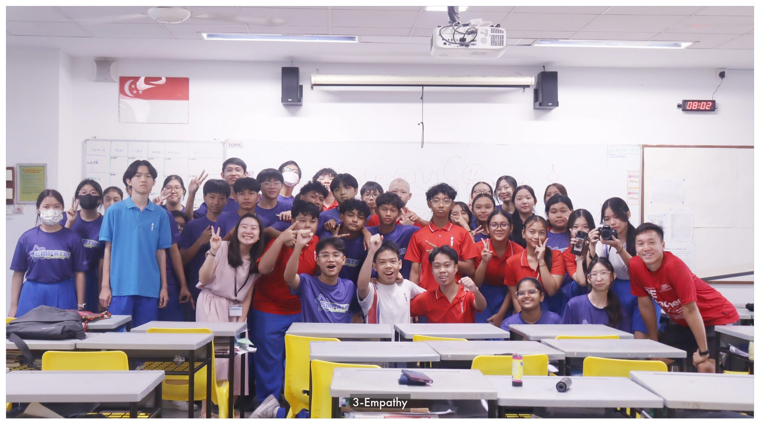 A large group of students and teachers pose together in a classroom with a Singapore flag on the wall.