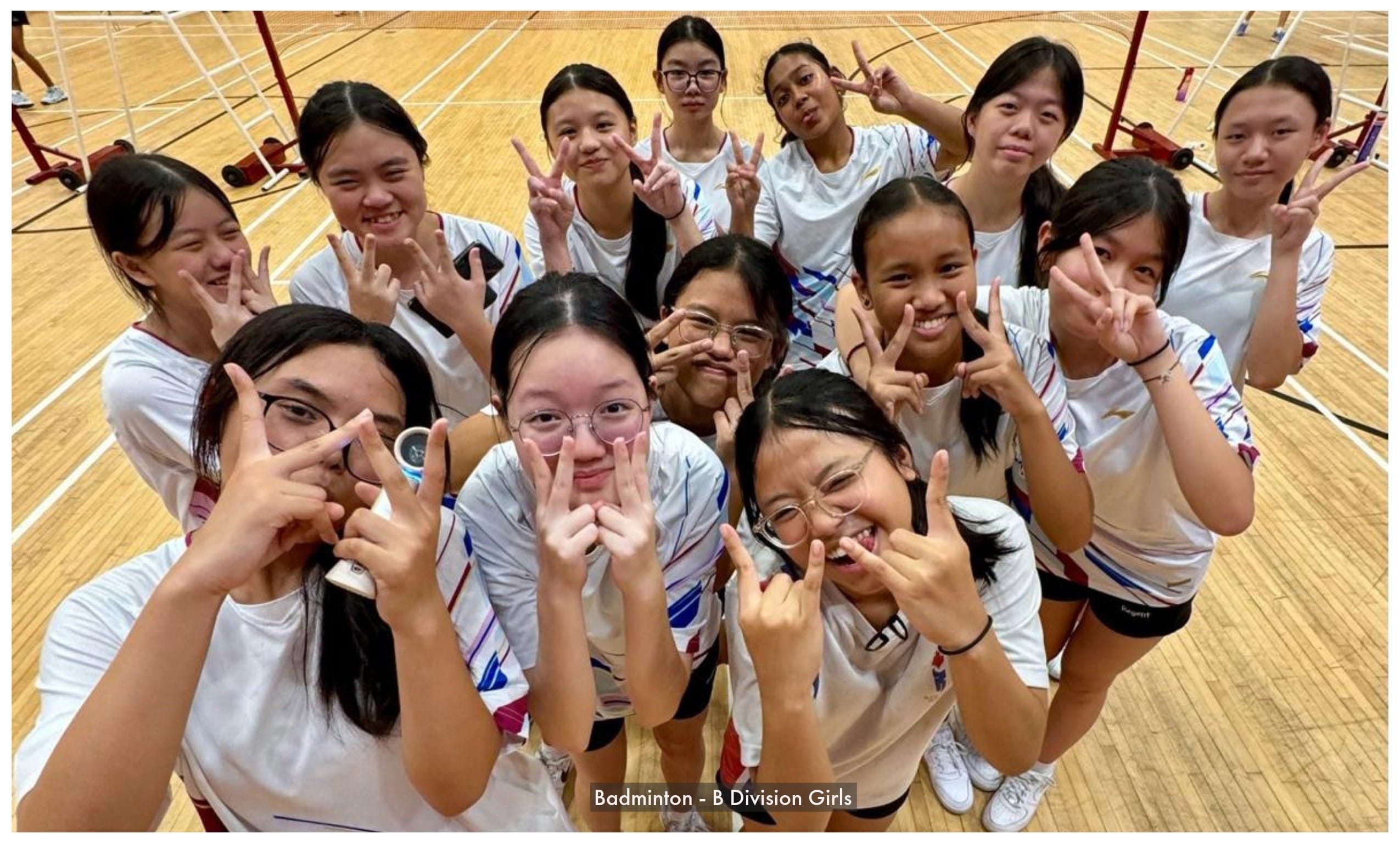 A group of teenage girls in white sports uniforms pose with peace signs on an indoor badminton court.