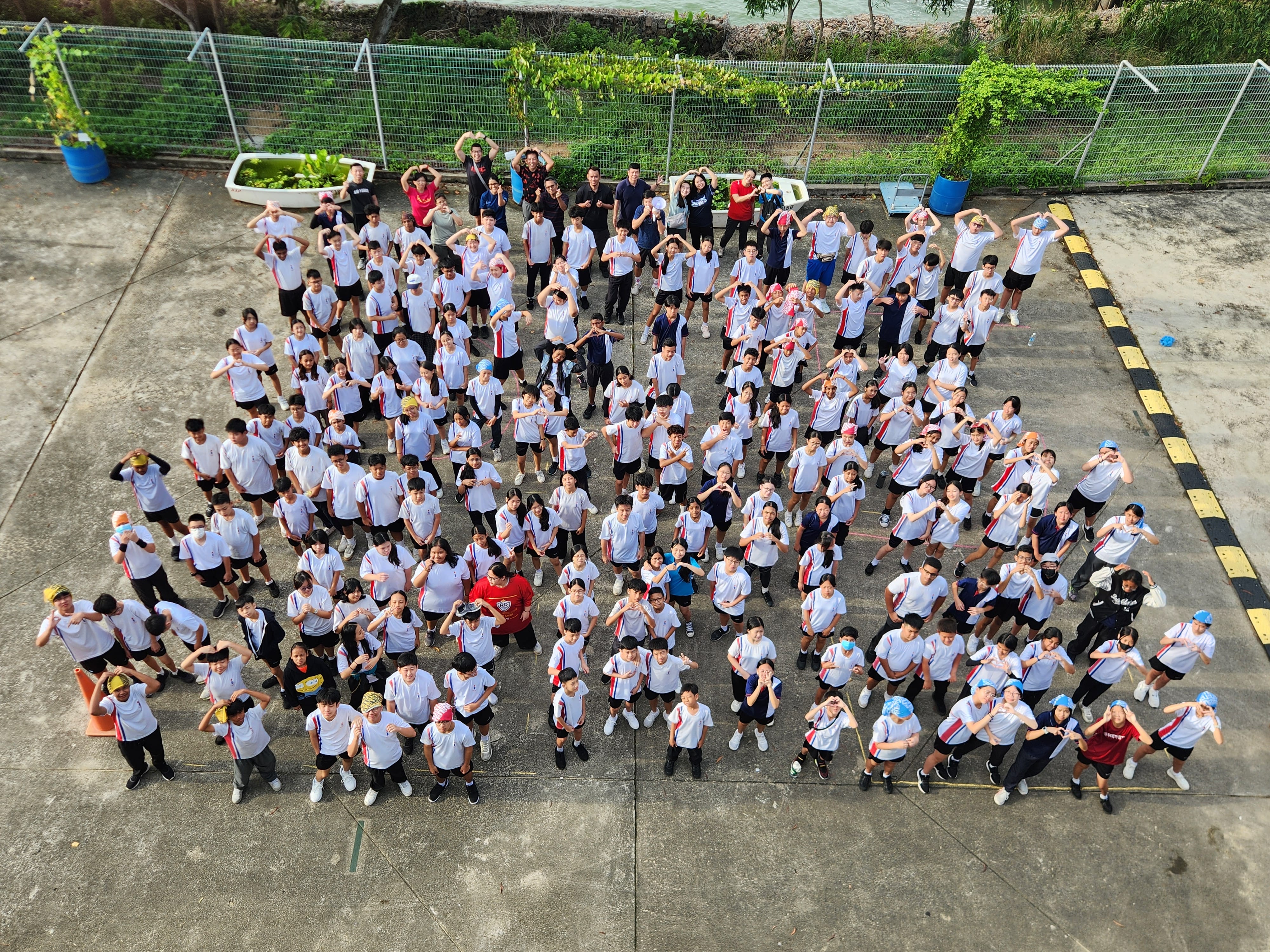 Overhead view of a large group in white shirts standing in a lot, many making heart shapes with arms.