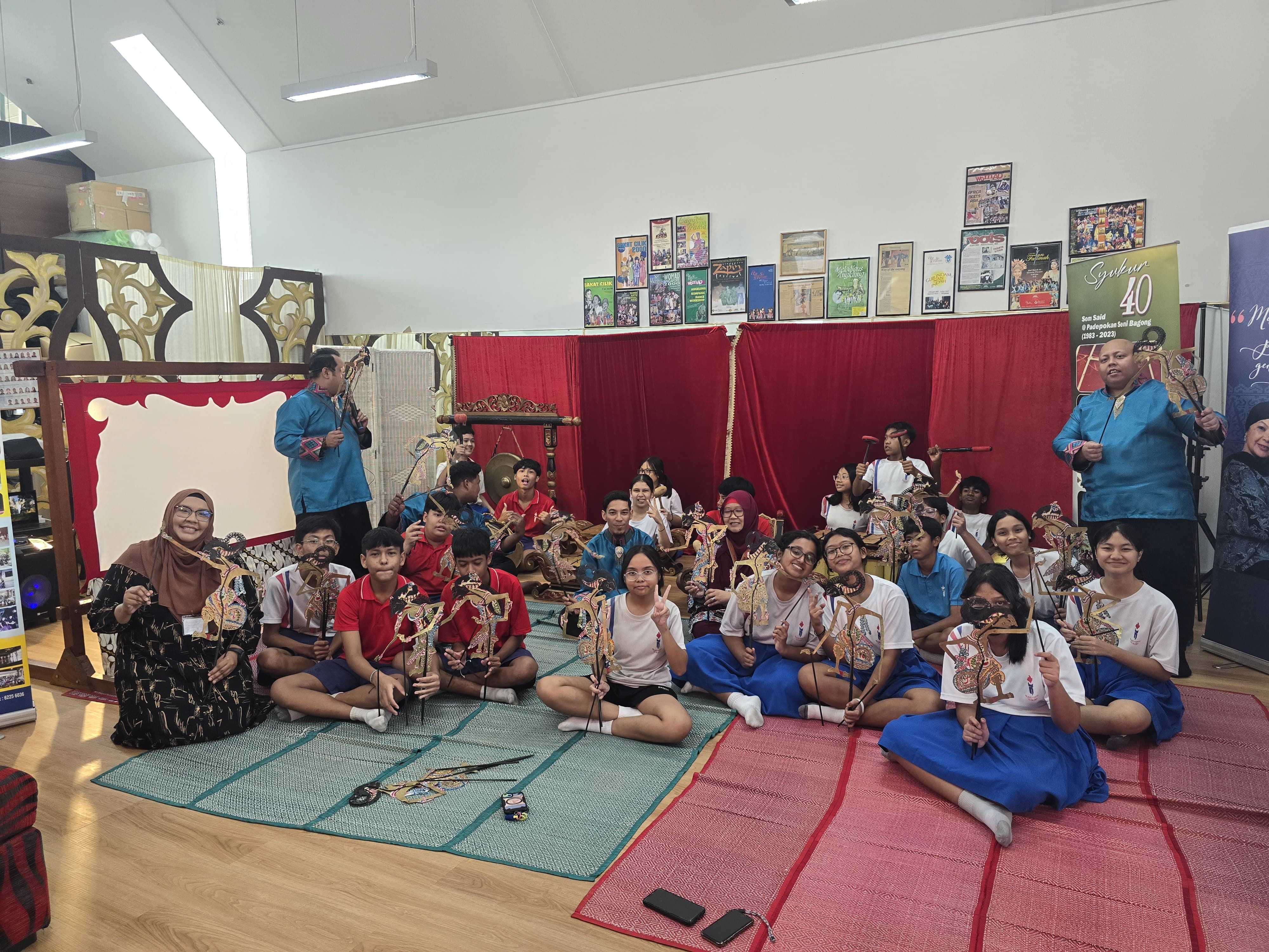 Students sit on mats holding traditional wayang puppet figures during a cultural workshop indoors.