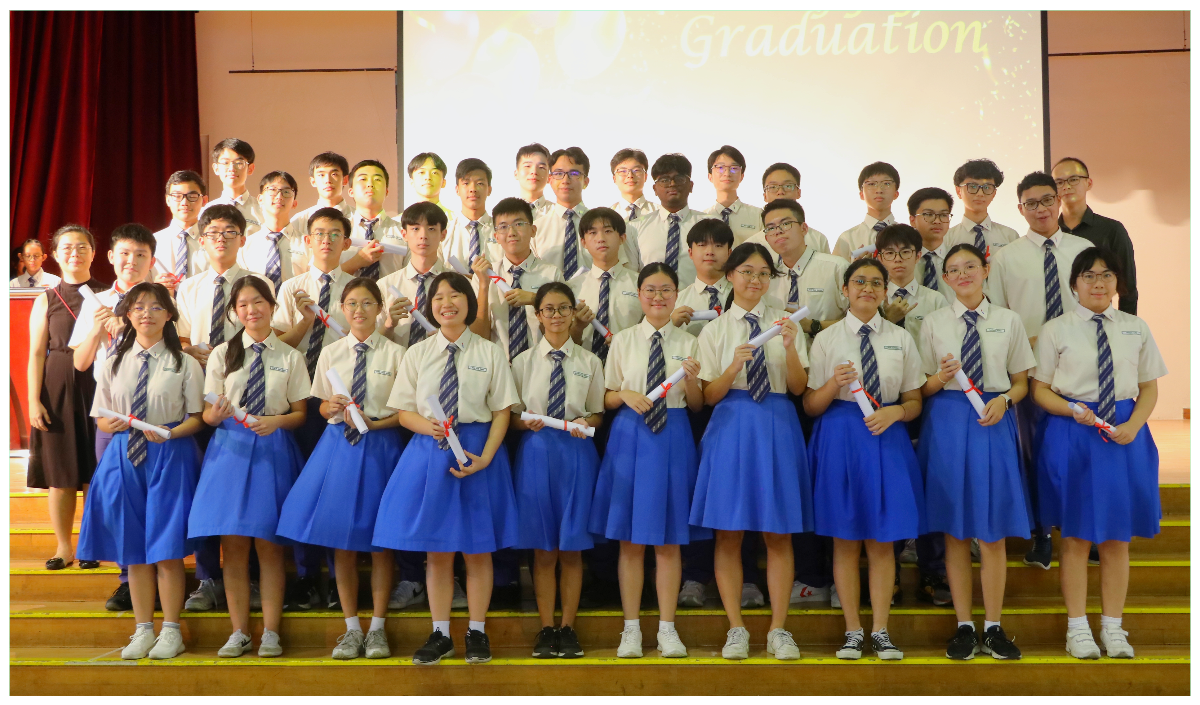 A large group of students in school uniforms holding diploma scrolls pose on stage at a graduation ceremony.