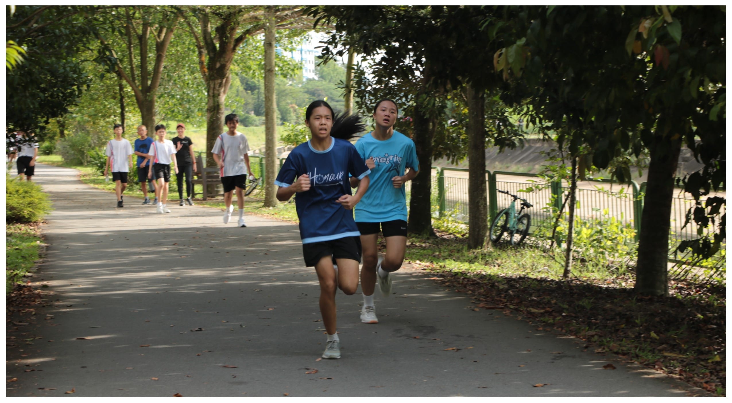 Two girls in blue shirts run along a tree-lined park path, with several other youth walking behind them.