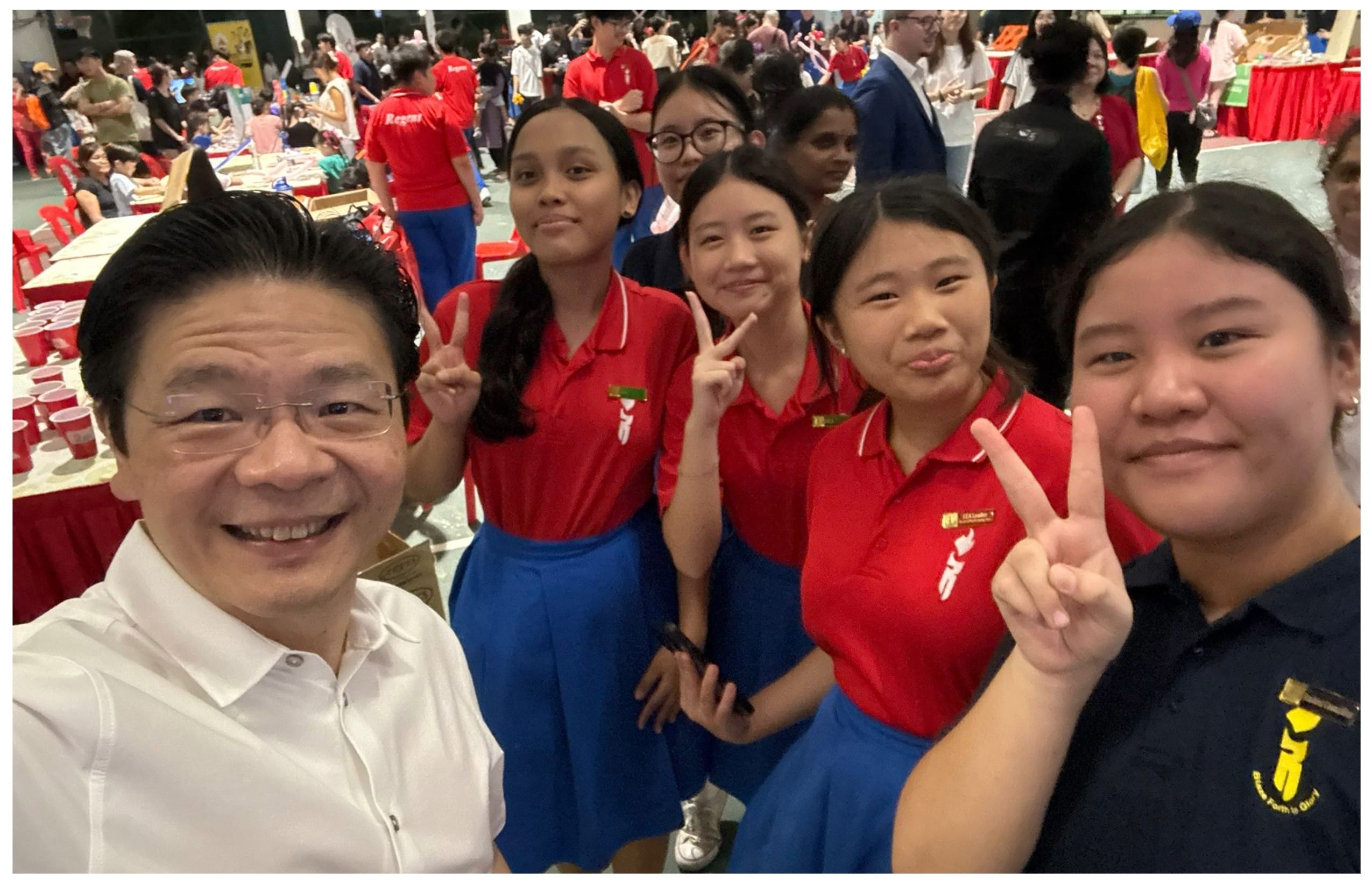 A man in a white shirt takes a selfie with four teenage girls in red and blue uniforms giving peace signs at a busy event.
