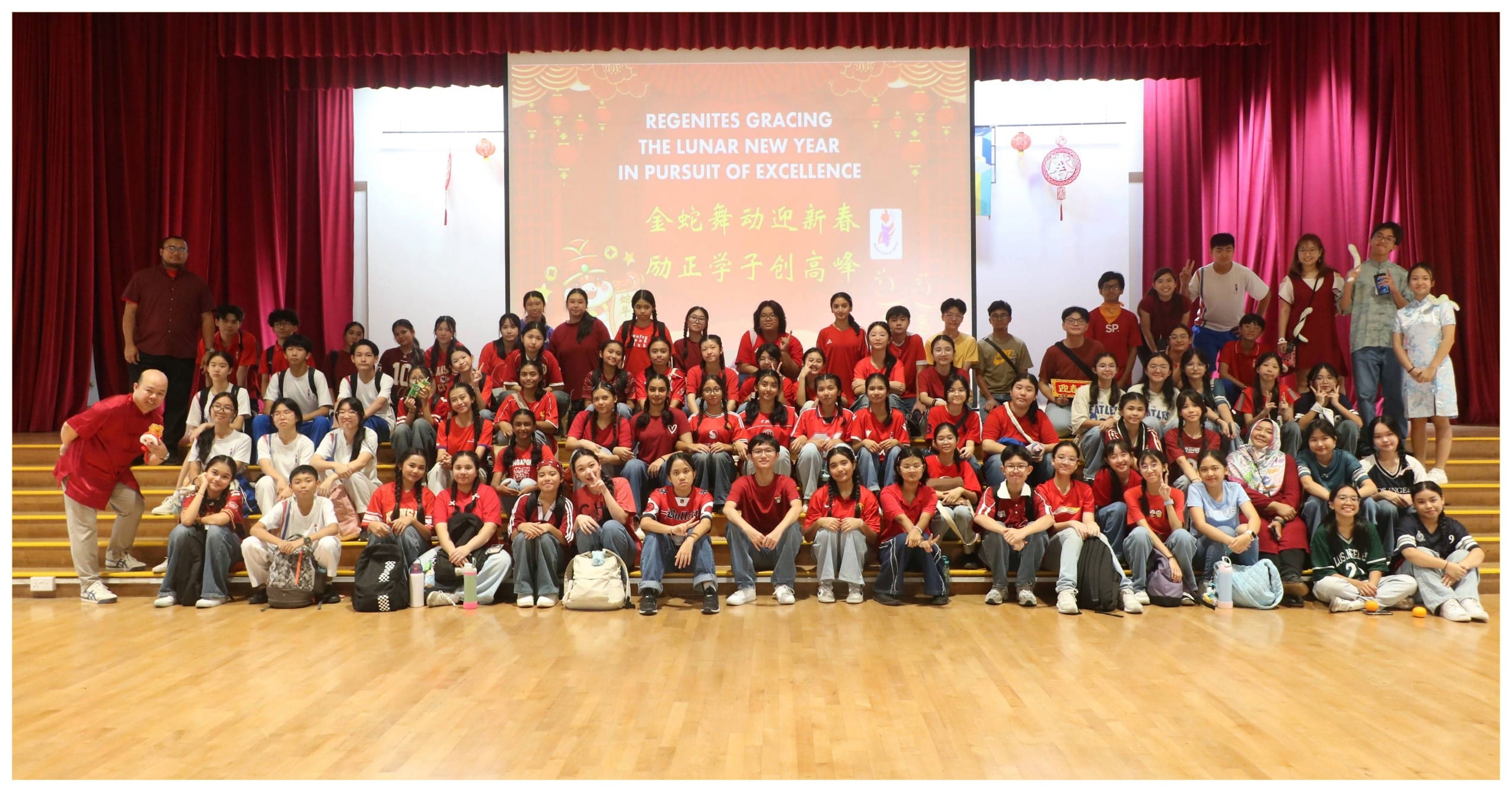 Large group of students in red shirts posing in a school hall before a Lunar New Year celebration banner.