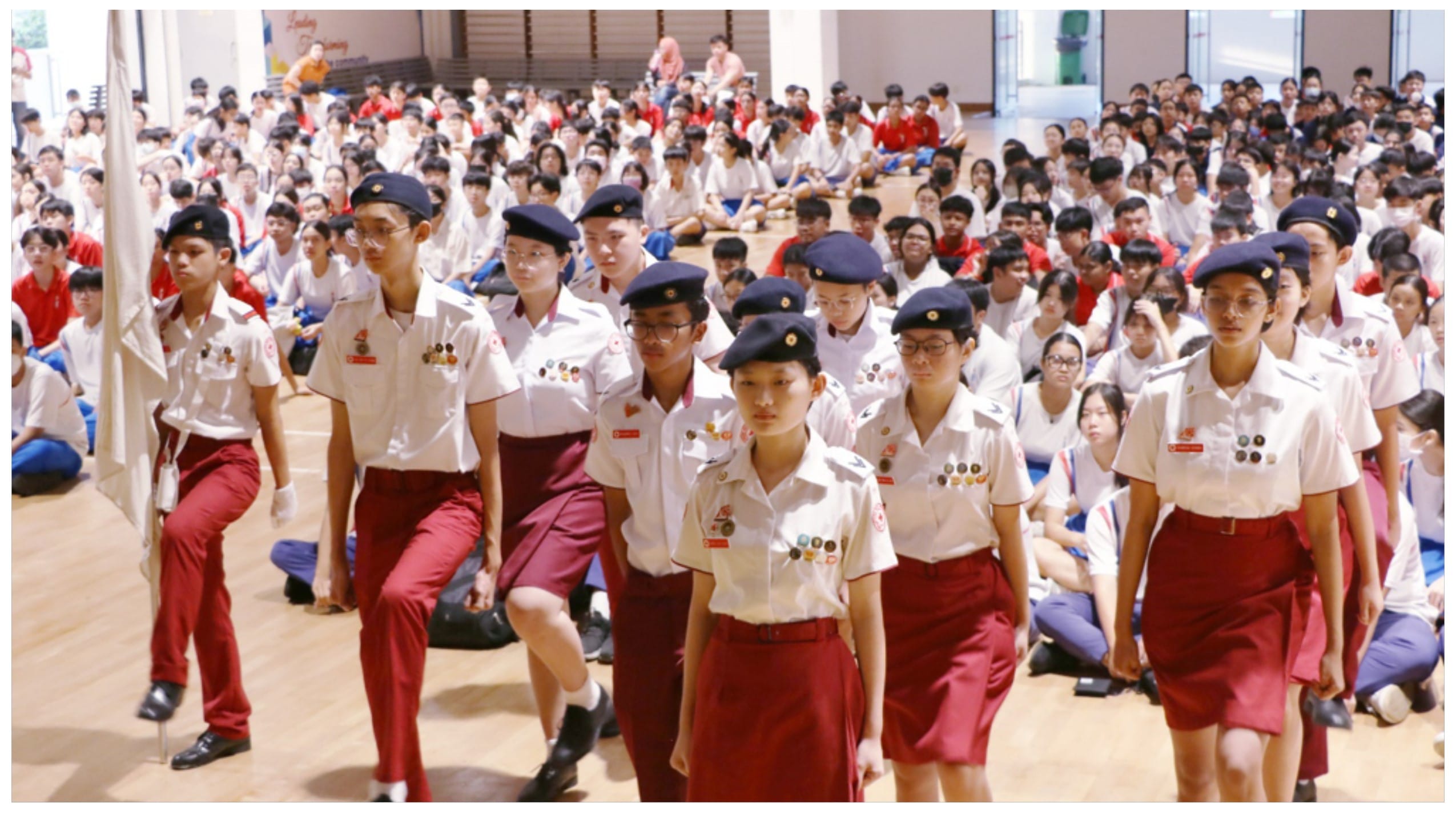 Students in white shirts, red bottoms, and navy berets march in a school hall while a large crowd of students sits watching.