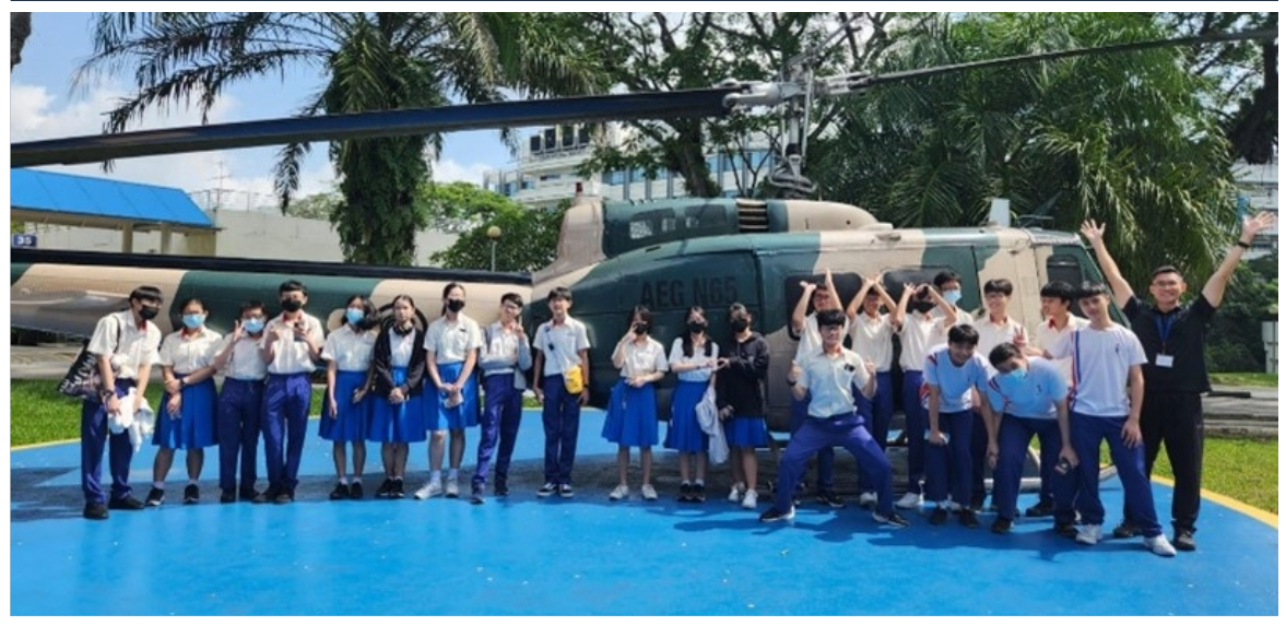 A group of uniformed students posing in front of a camouflage military helicopter on a blue outdoor pad.