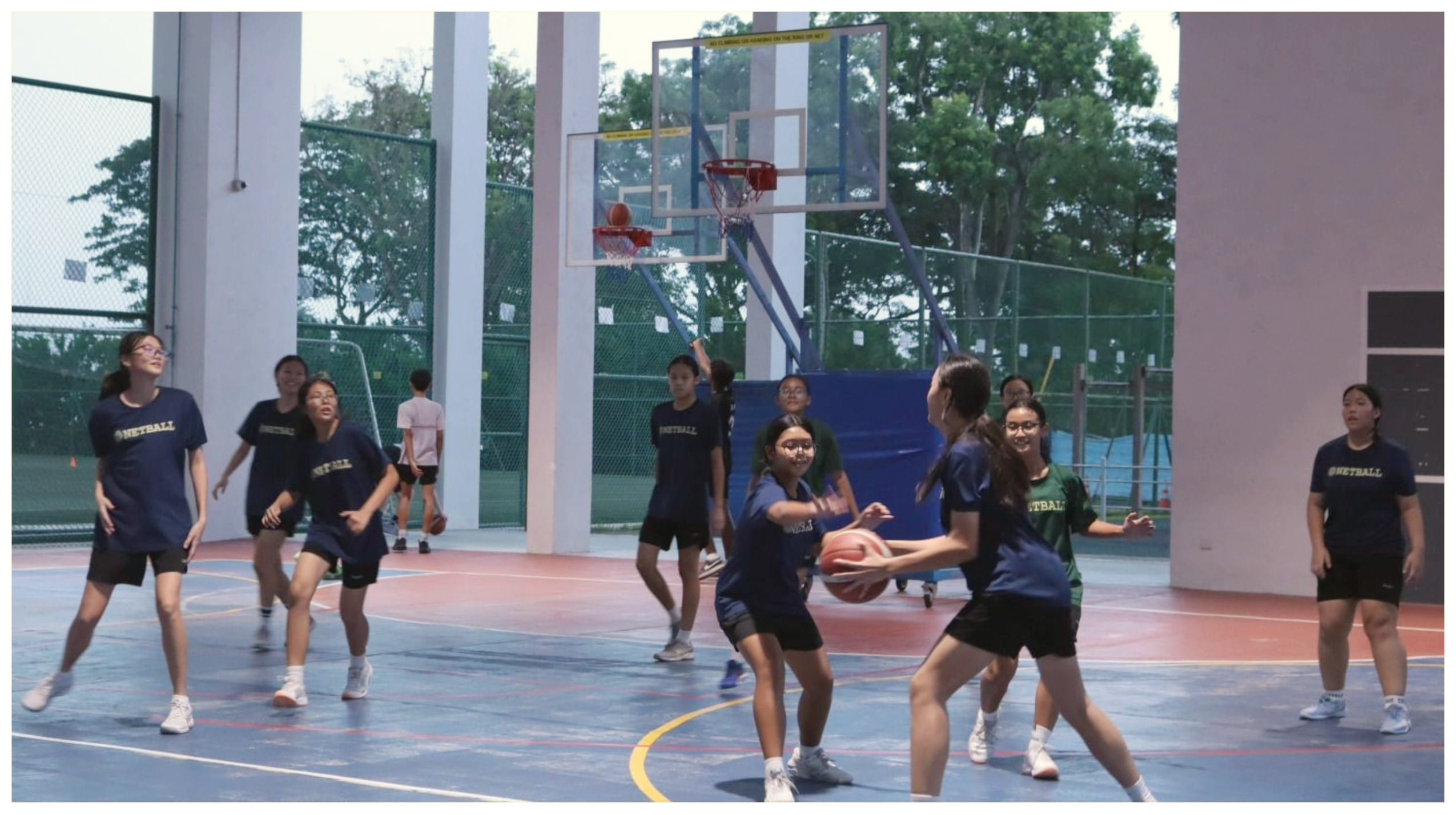 Girls in navy netball shirts play basketball on an outdoor court, with one player holding the ball near the hoop.