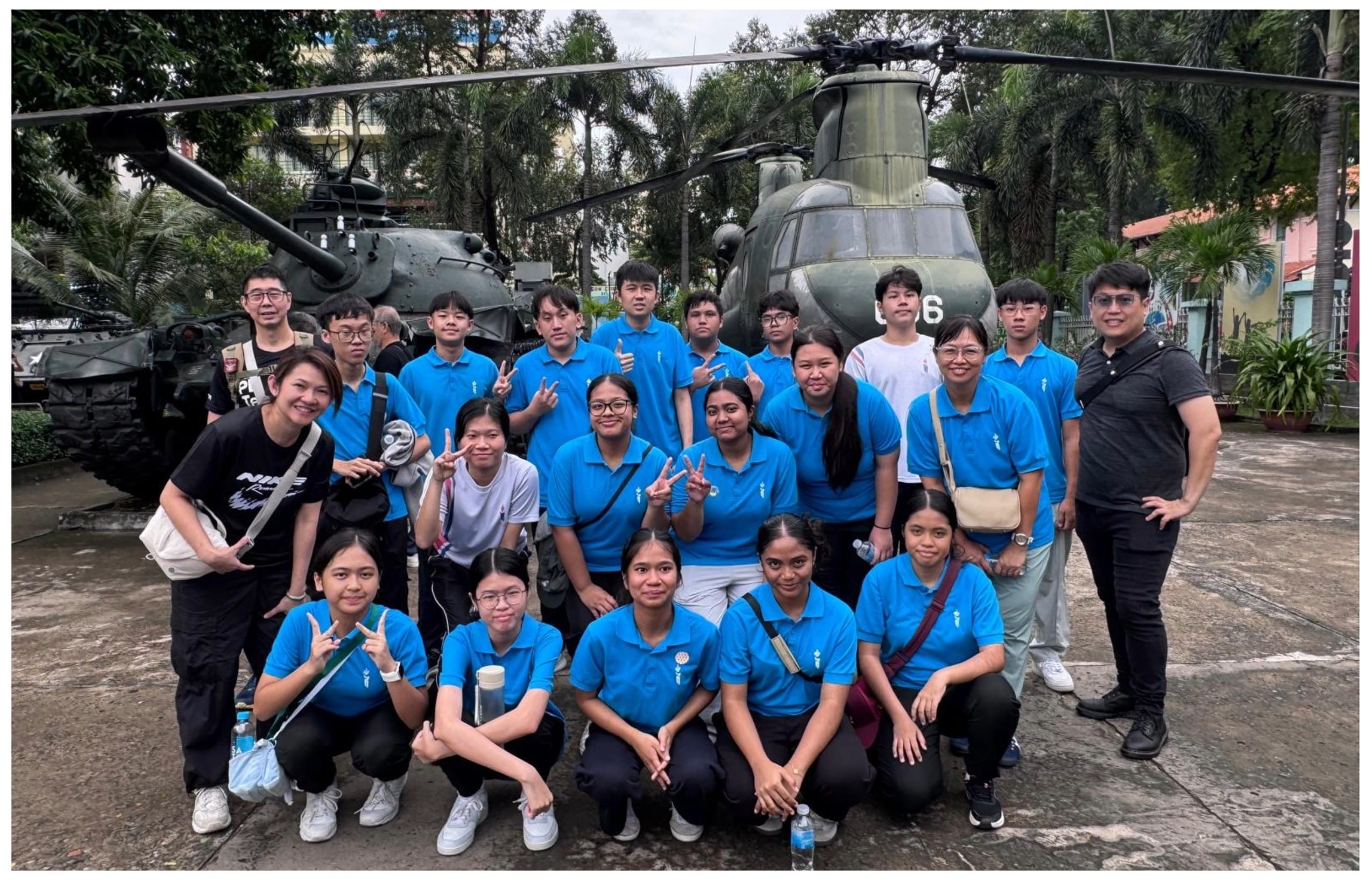 A group of young people in blue polo shirts pose in front of a military helicopter and tank outdoors.
