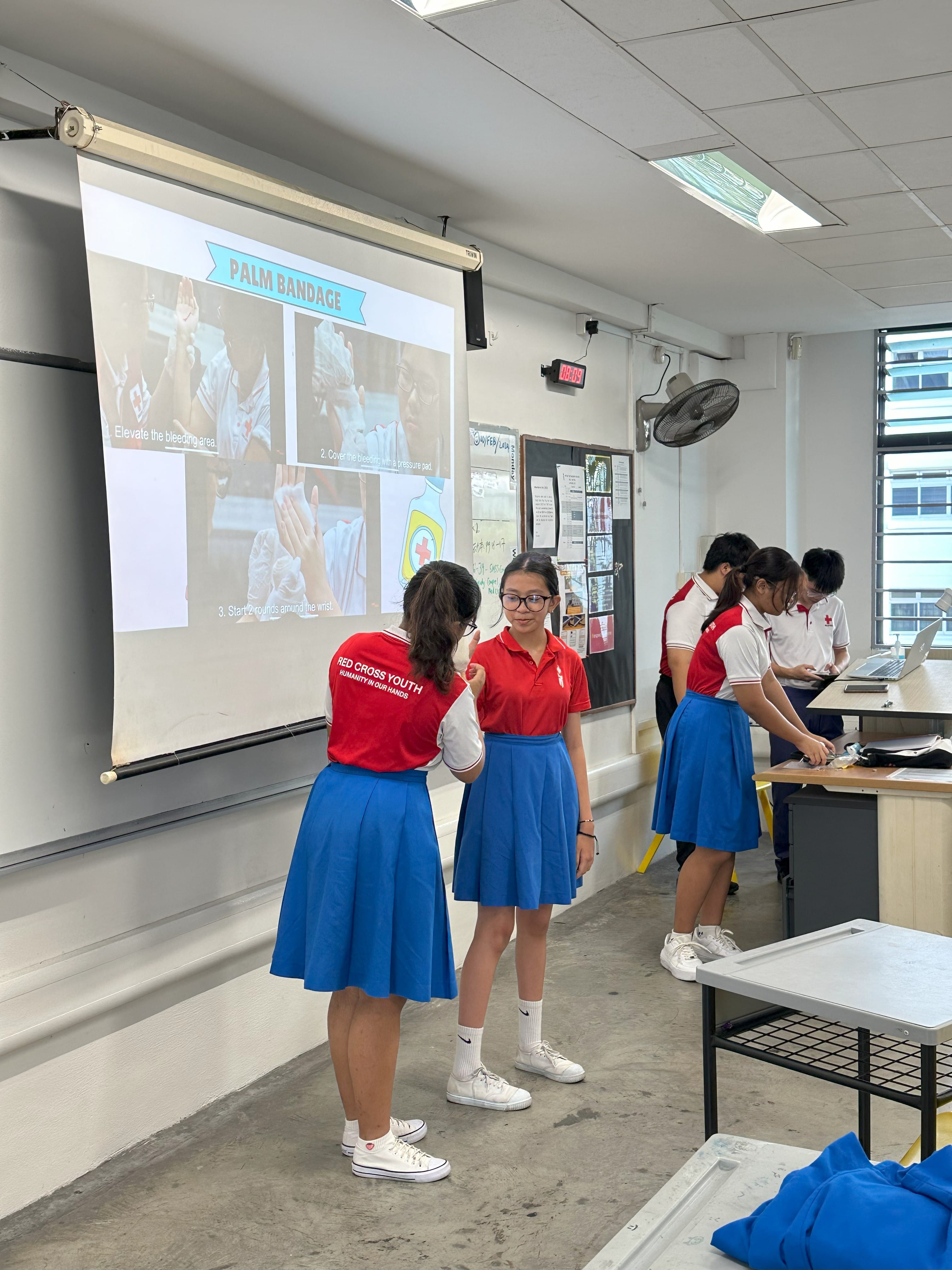 Red Cross Youth students in red shirts practice palm bandaging in a classroom with a first aid slideshow projected.