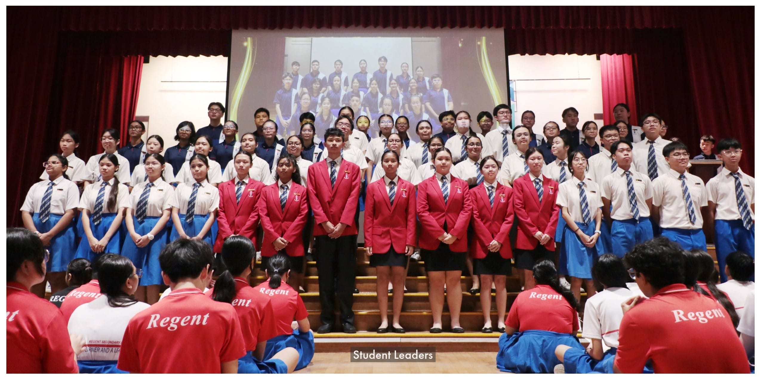 Large group of Regent Secondary school students in uniform pose on stage, with student leaders in red blazers at front.