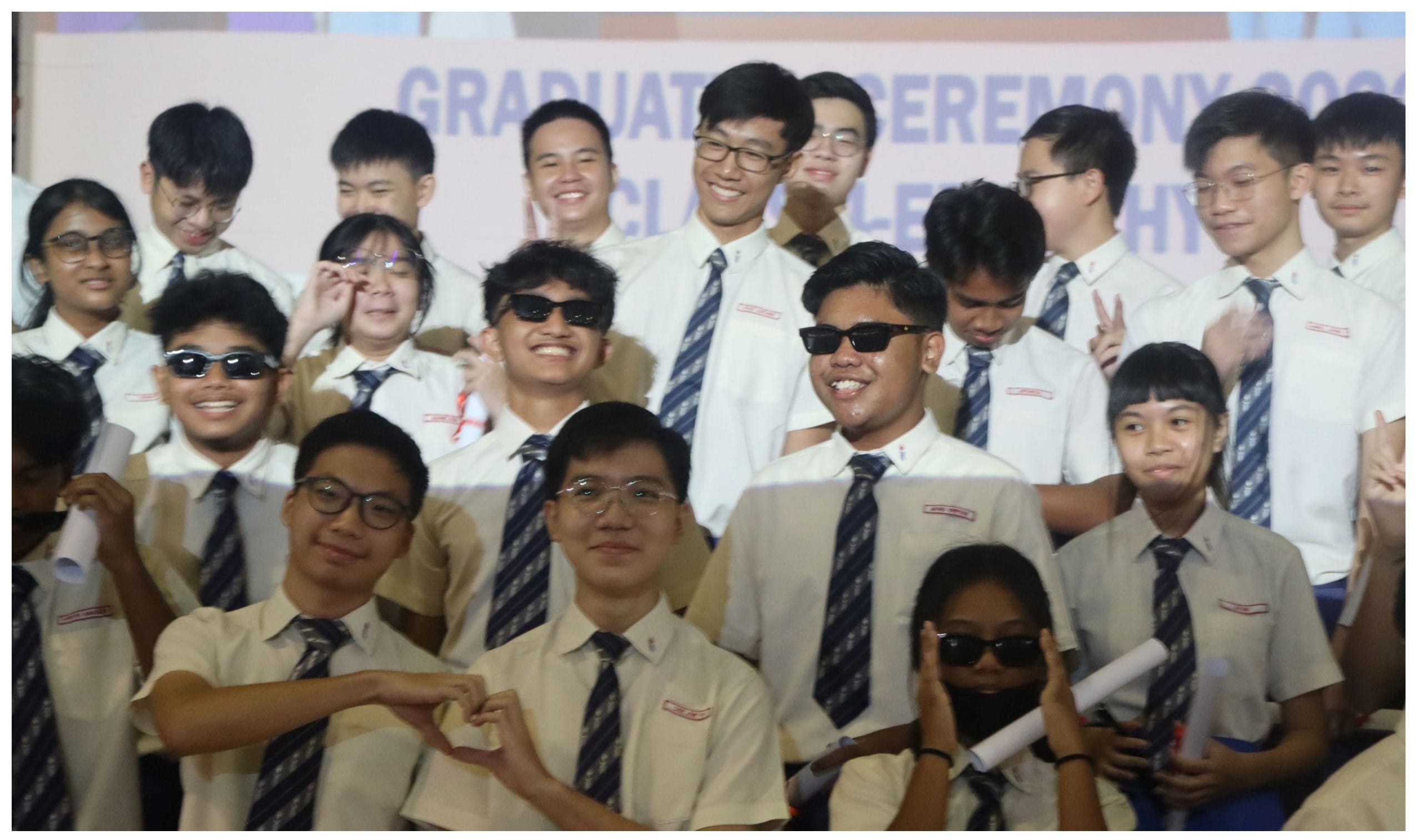 Group of students in uniforms with ties posing and smiling at a graduation ceremony backdrop.
