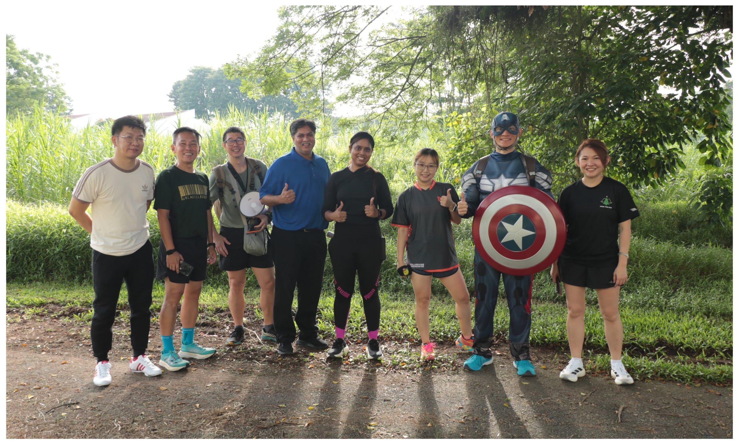 Seven people pose outdoors; one wears a Captain America costume with shield, others in casual sportswear, surrounded by greenery.