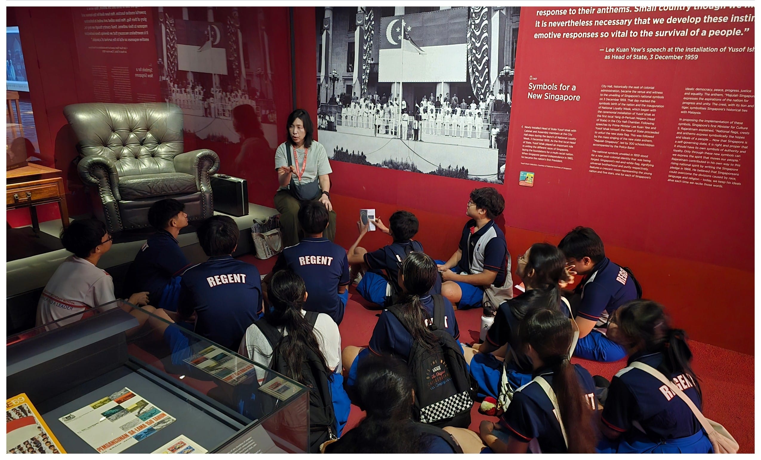 A guide speaks to students in blue Regent uniforms seated on the floor of a Singapore history museum exhibit.