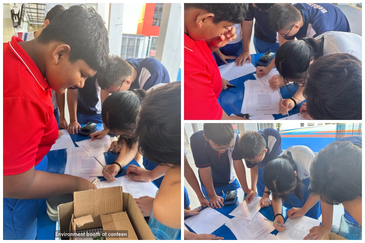 Students in blue and red uniforms huddle together writing on paper sheets at an environment booth in a canteen.