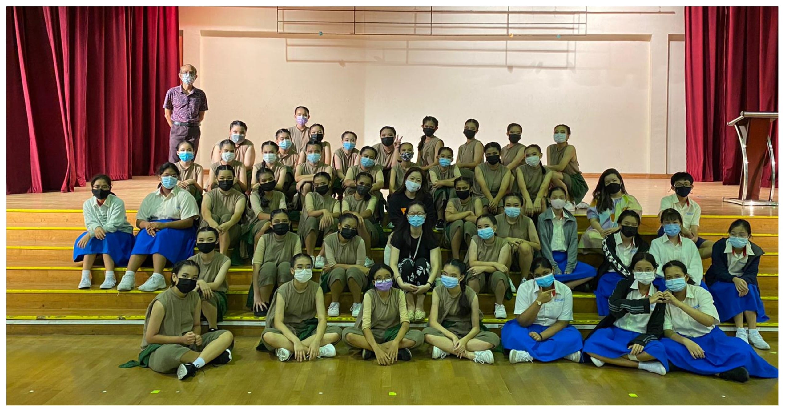 Large group of masked students and adults posing on bleachers in a school hall with red curtains.
