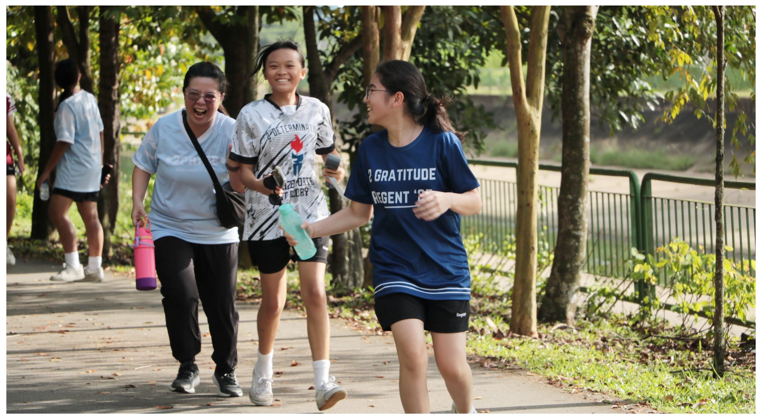Three smiling women jogging along a tree-lined park path, carrying water bottles, wearing casual sportswear.