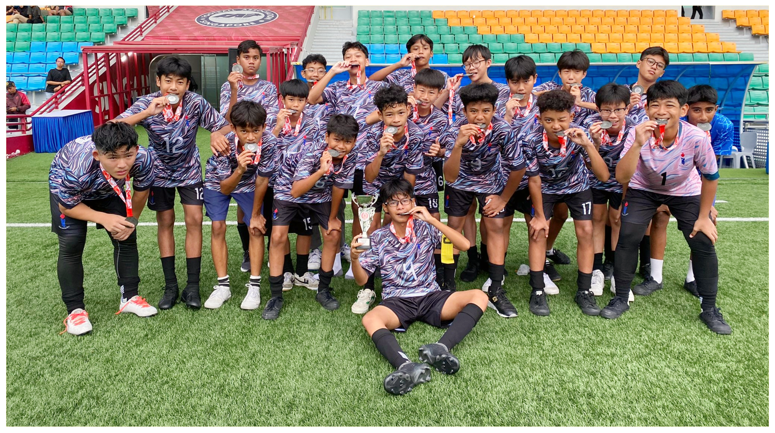 Youth soccer team poses on turf, wearing medals; one sits holding a trophy, stadium seats behind.