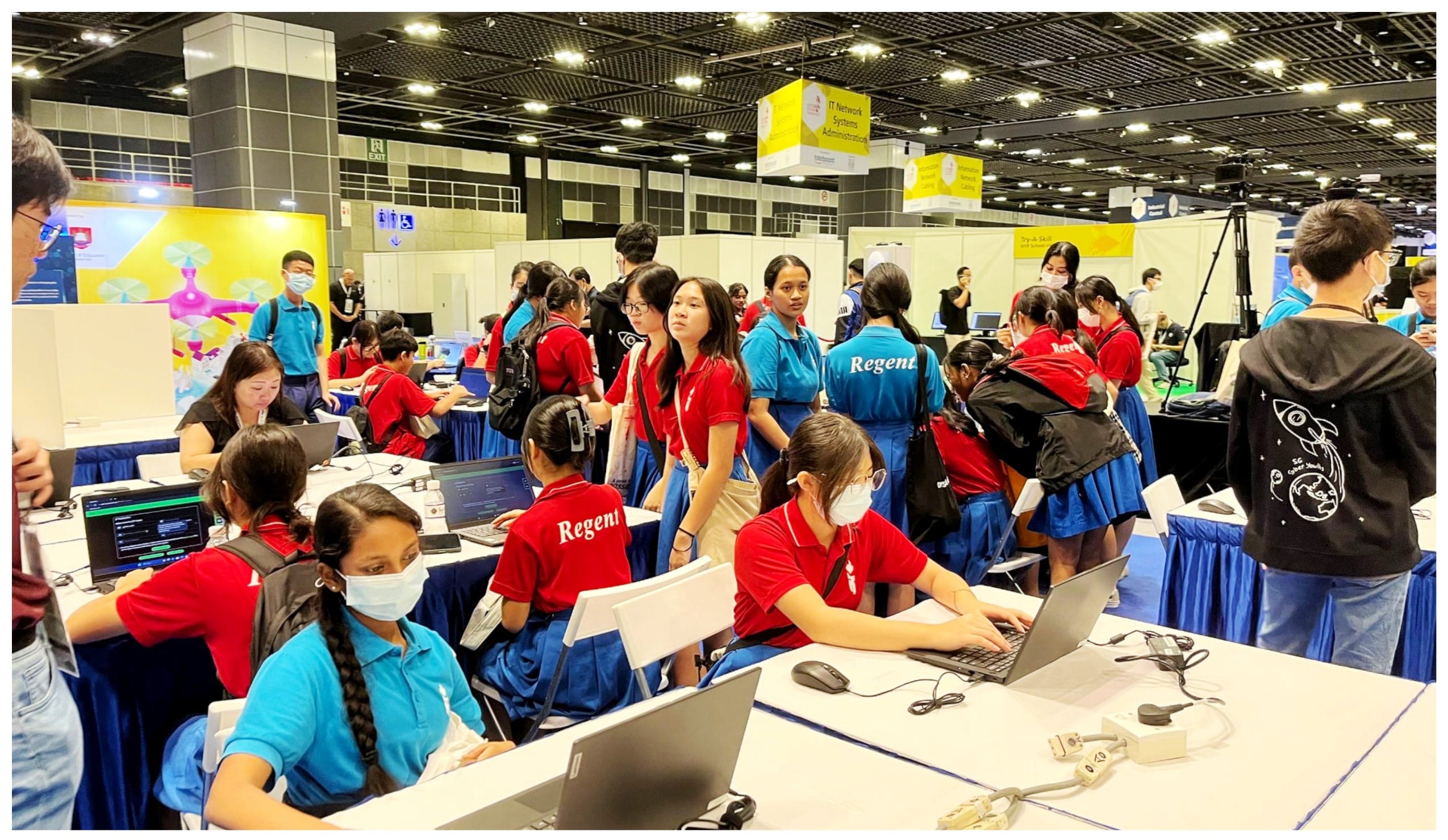 Students in red and blue uniforms work on laptops at tables inside a large convention hall.