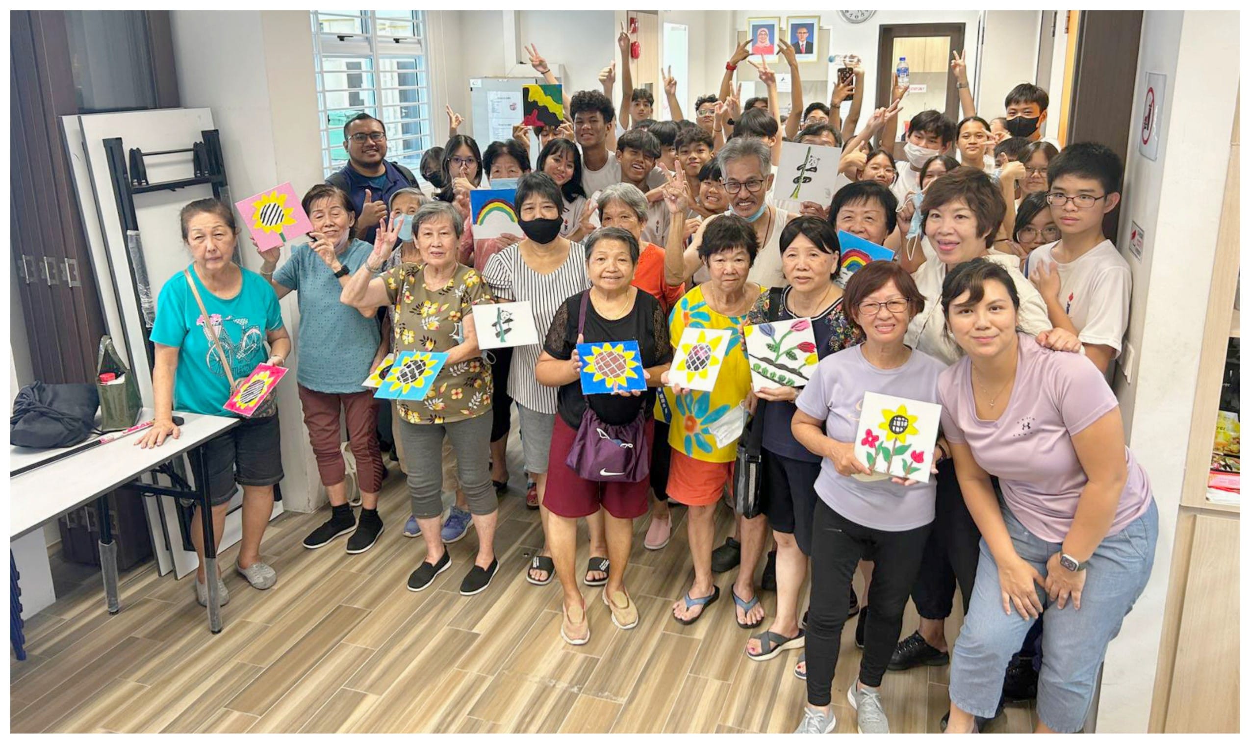 A large group of adults and teens smiling indoors, holding up colorful painted canvases with flowers and rainbows.
