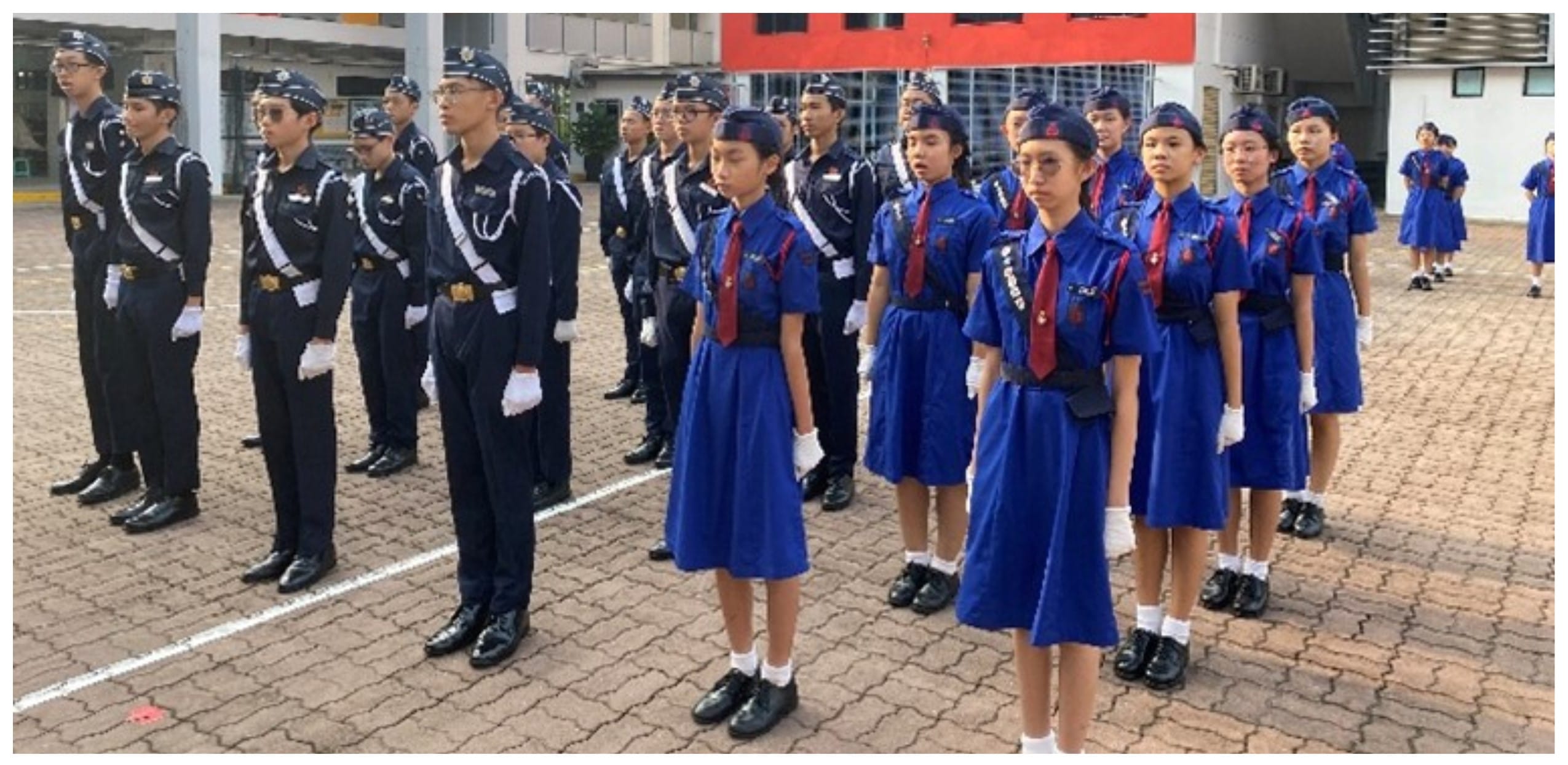 Students in dark and blue uniforms with caps stand at attention in rows on a paved courtyard during a parade.