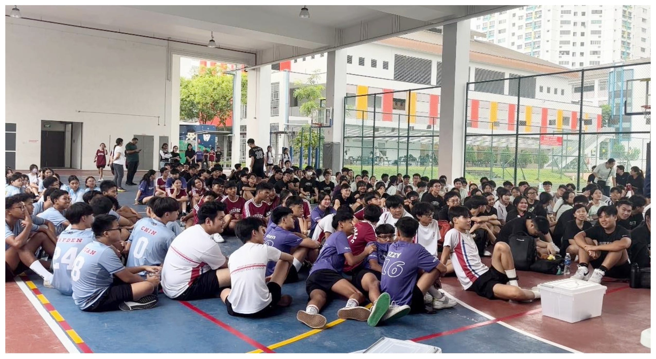 Large group of students in sports jerseys sitting on an outdoor covered basketball court at a school.