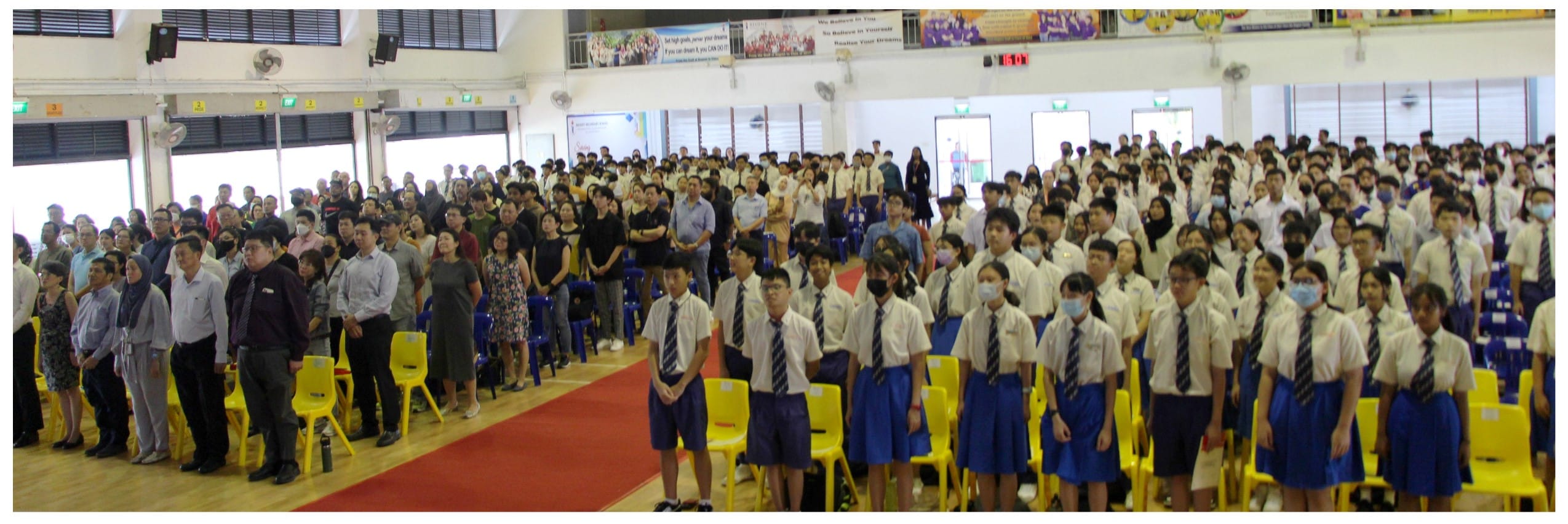Students in white shirts and blue uniforms stand in rows beside adults in a school hall with yellow chairs.