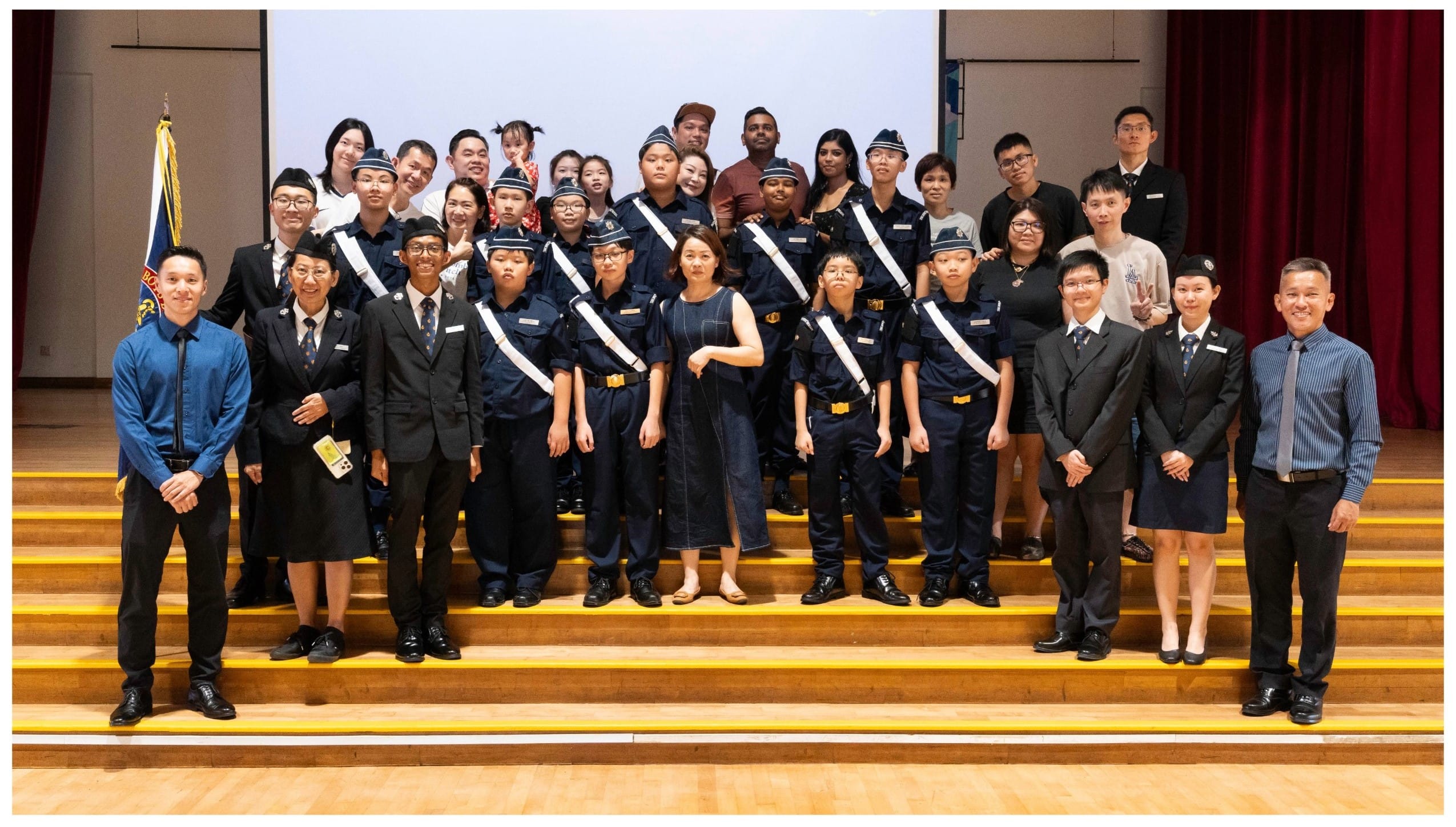 A large group of youth in navy uniforms with white sashes pose on stage steps with adults in formal attire.