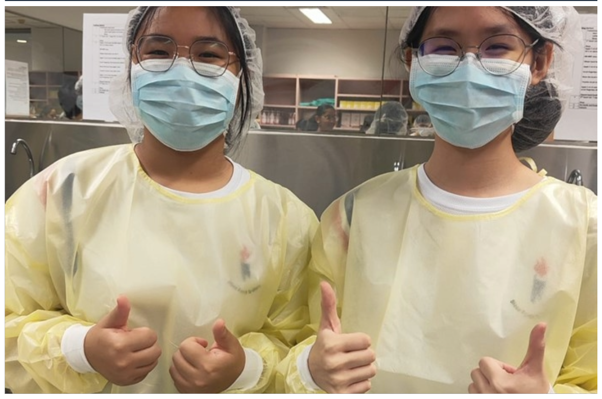 Two young women in yellow gowns, blue masks, hair nets, and glasses give thumbs up in a medical setting.