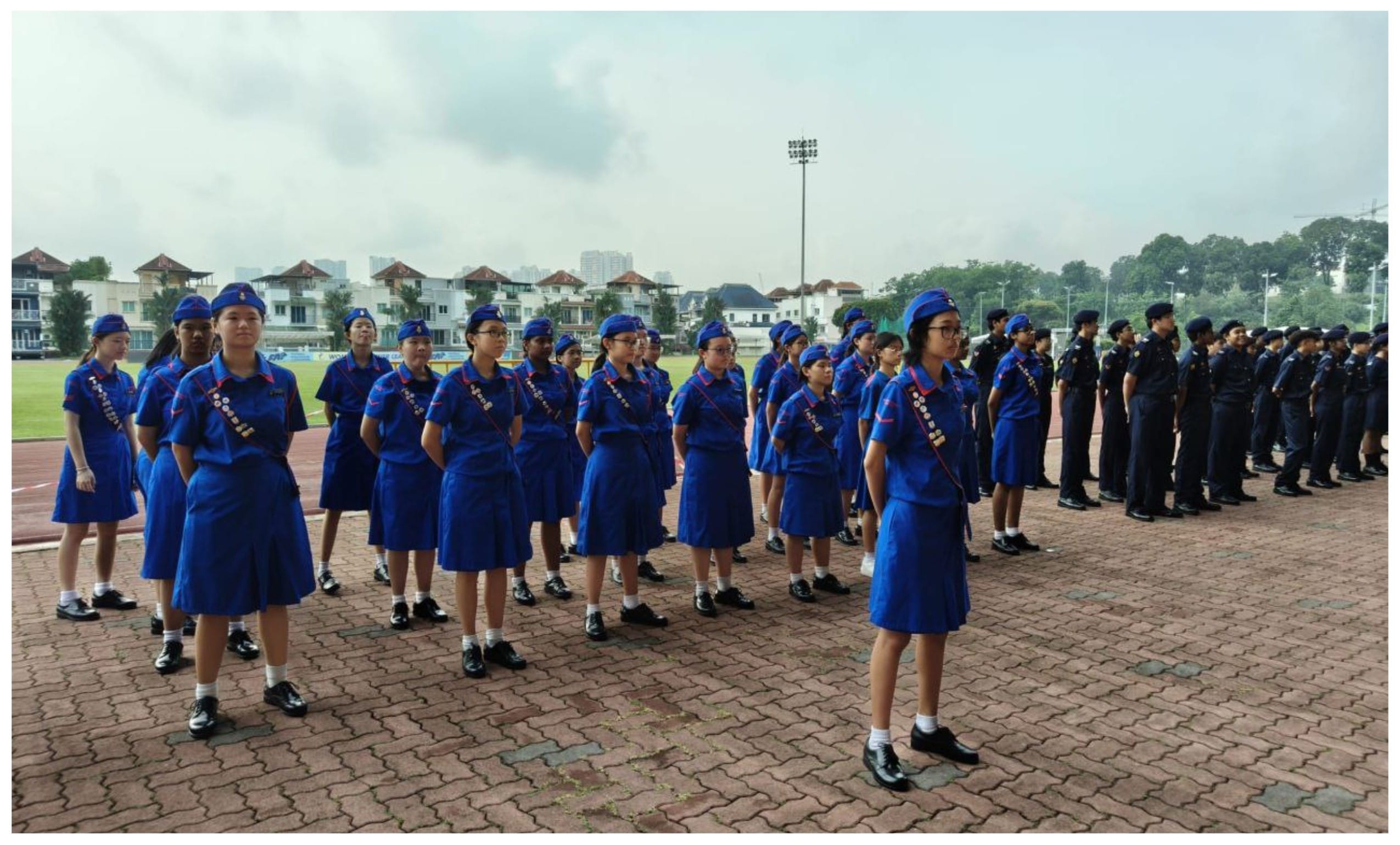 Young people in blue uniforms and caps stand in formation on a paved area near a sports field with buildings behind them.