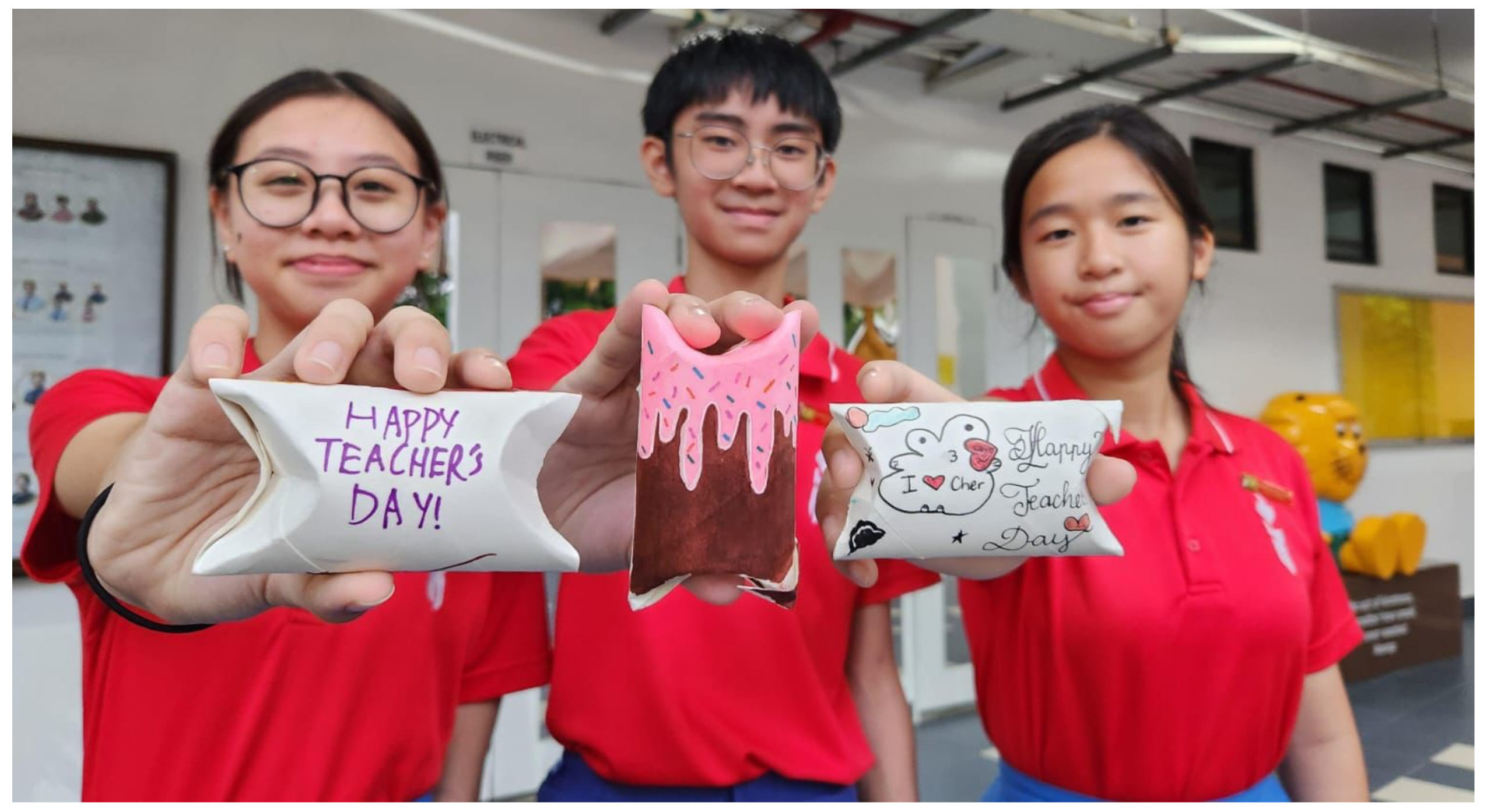 Three students in red uniforms hold hand-decorated Teacher's Day gifts toward the camera, smiling.