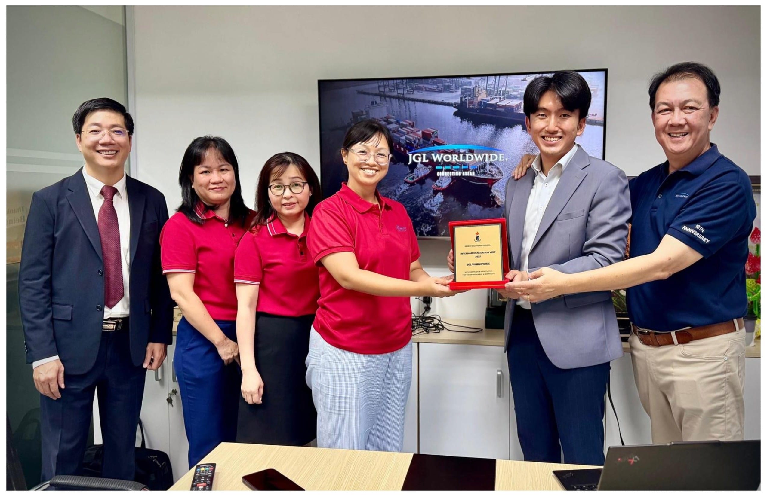 Six people smile indoors as two hold a plaque for JGL Worldwide, with a screen displaying the company name behind them.