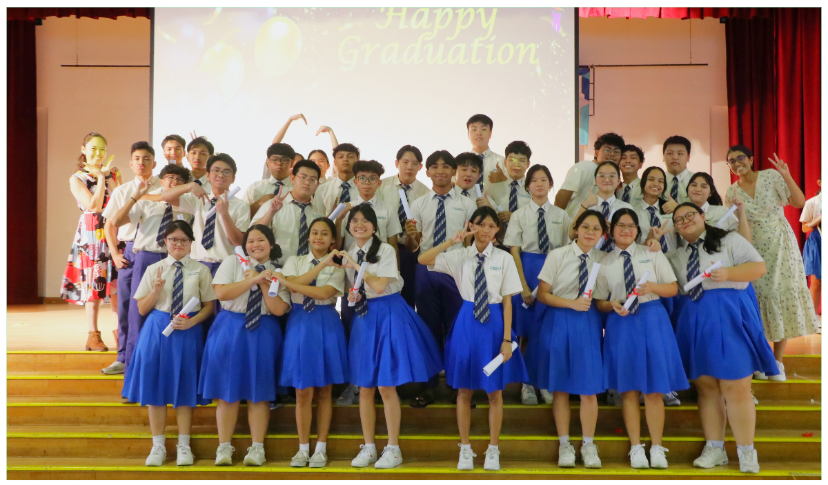 Students in school uniforms pose on stage steps holding diplomas, with a Happy Graduation screen behind them.