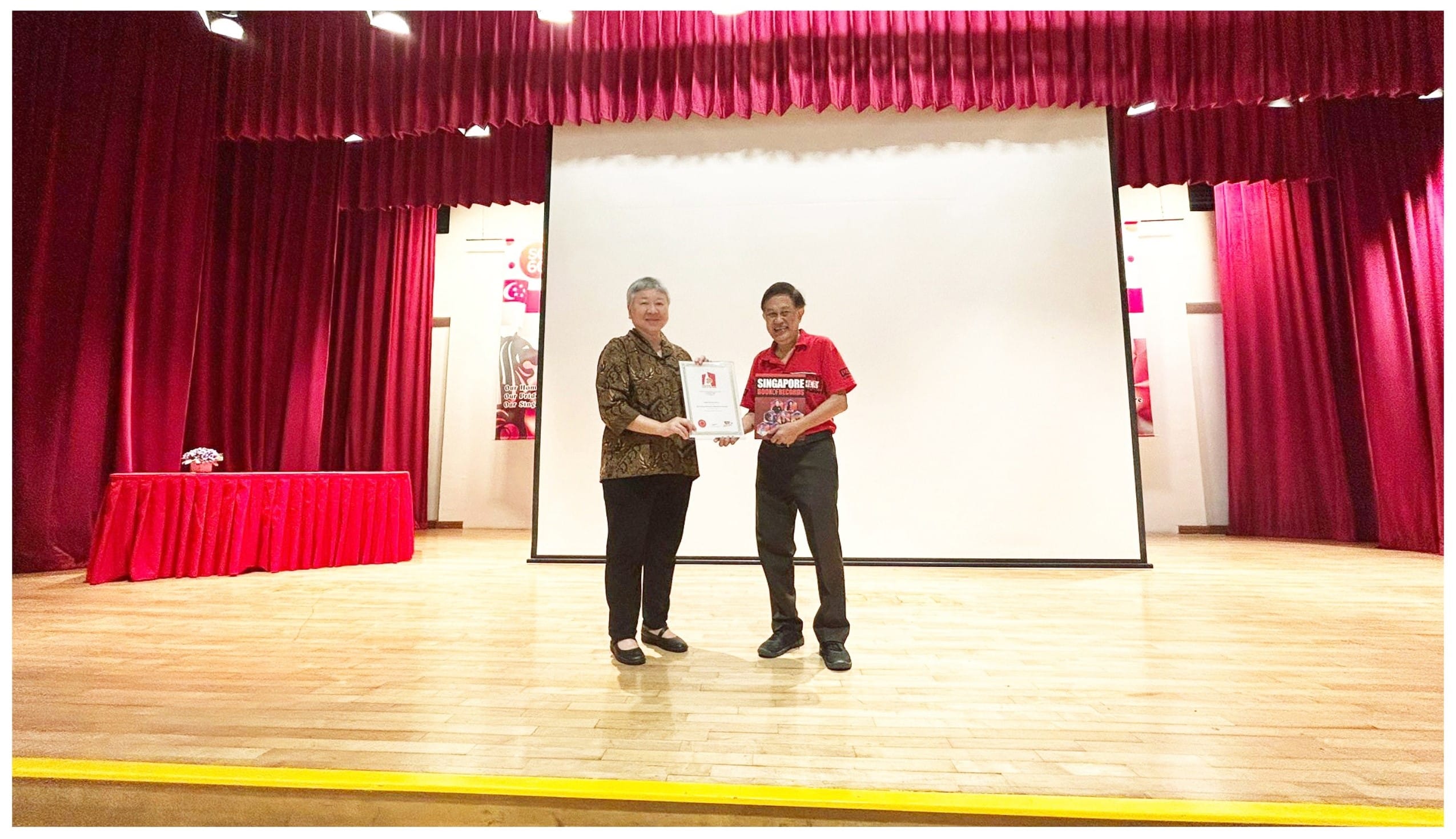 Two people stand on a wooden stage with red curtains, jointly holding a framed certificate between them.