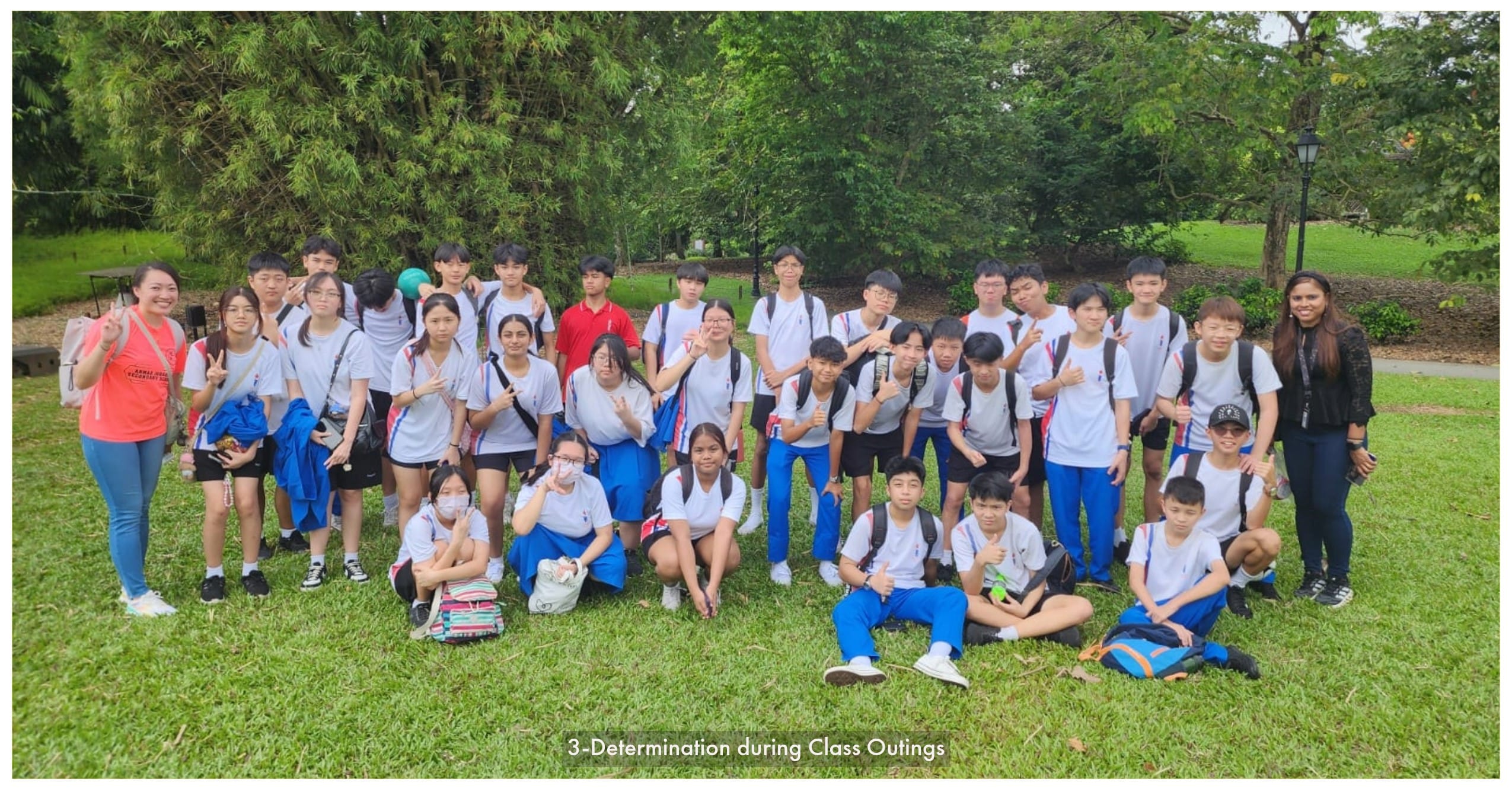 A large group of students in white and blue uniforms pose with two teachers on a grassy area surrounded by trees.