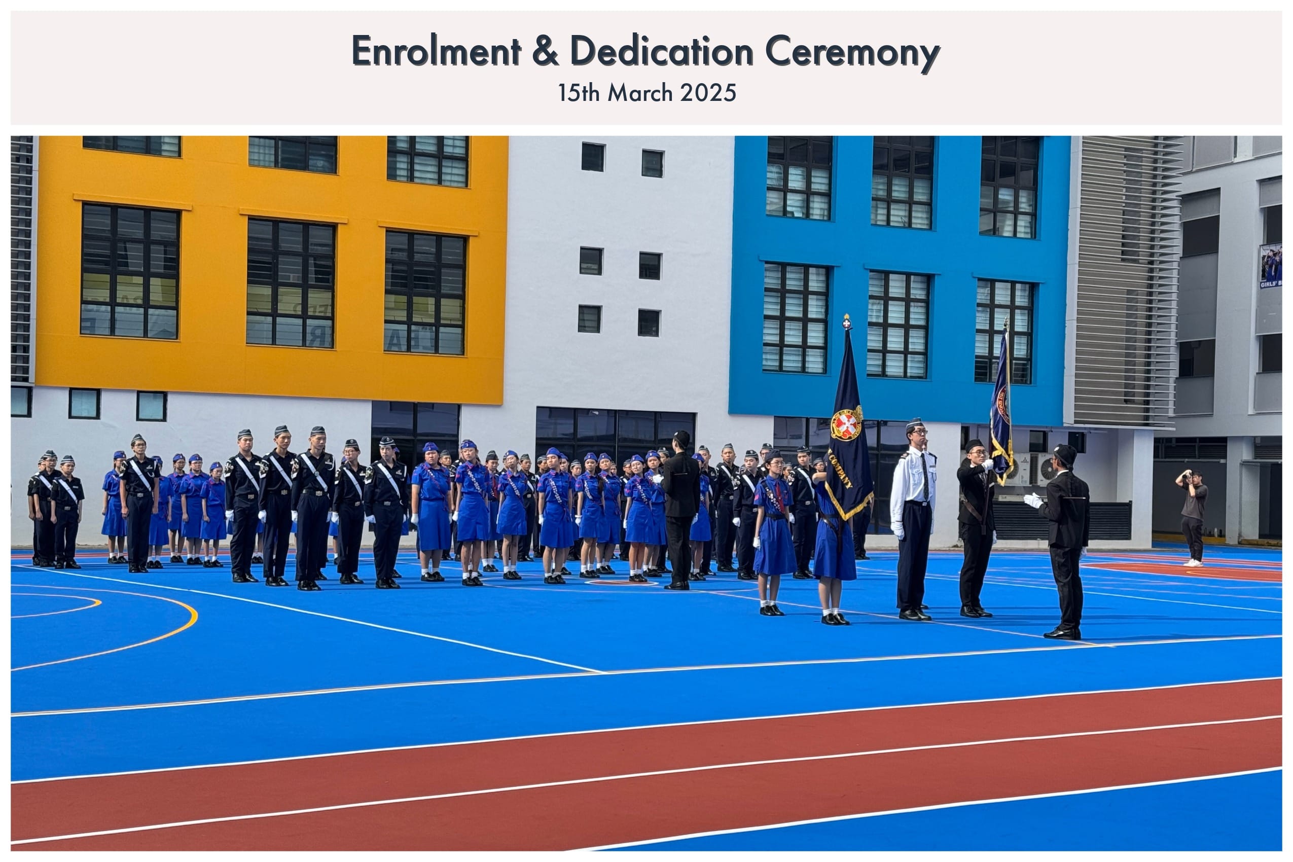 Students in blue and dark uniforms stand in formation on a colorful school court during an Enrolment & Dedication Ceremony.