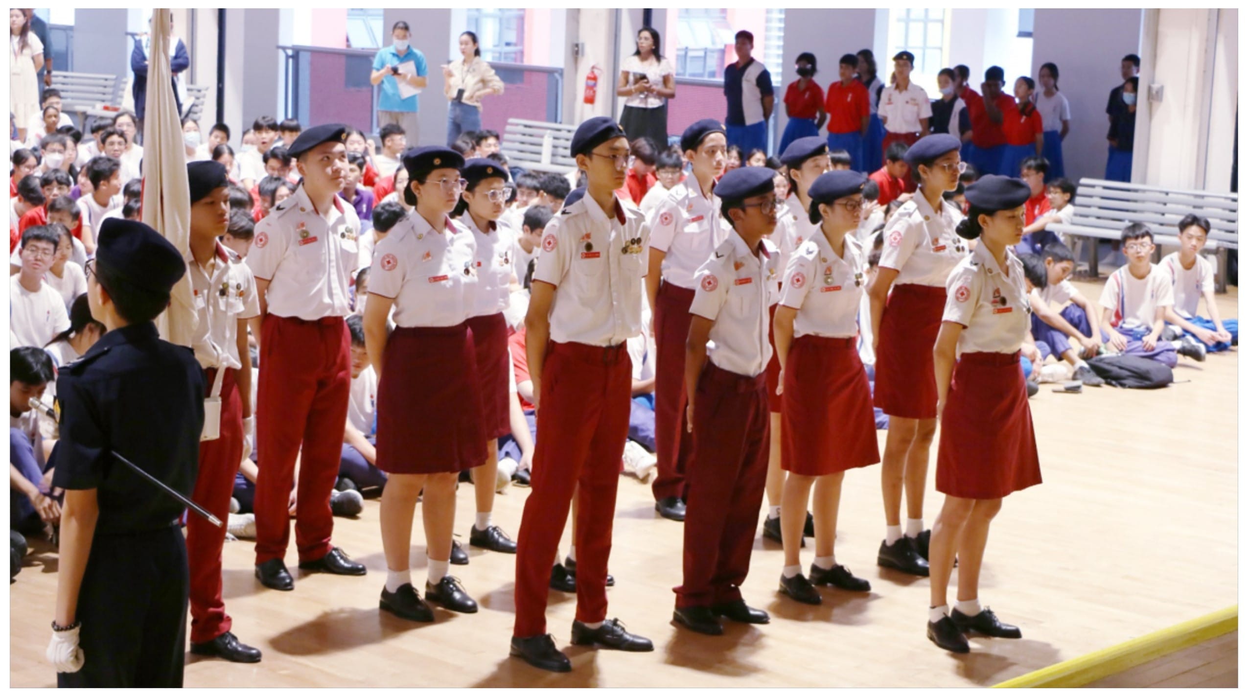 Students in white shirts, red bottoms, and dark berets stand in formation during a school ceremony in an indoor hall.