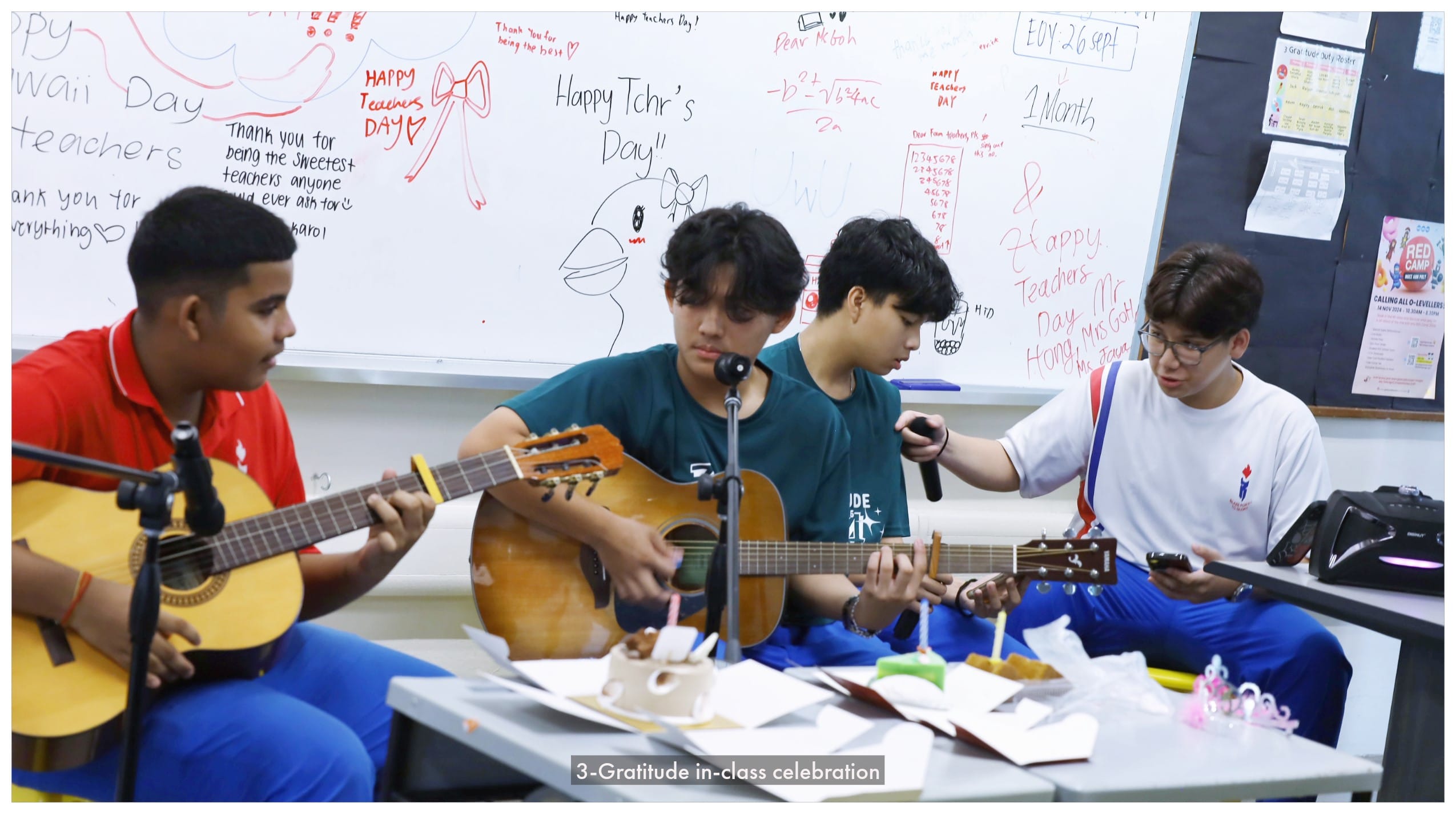 Four students play guitars and sing in a classroom decorated with Happy Teachers Day messages on a whiteboard.