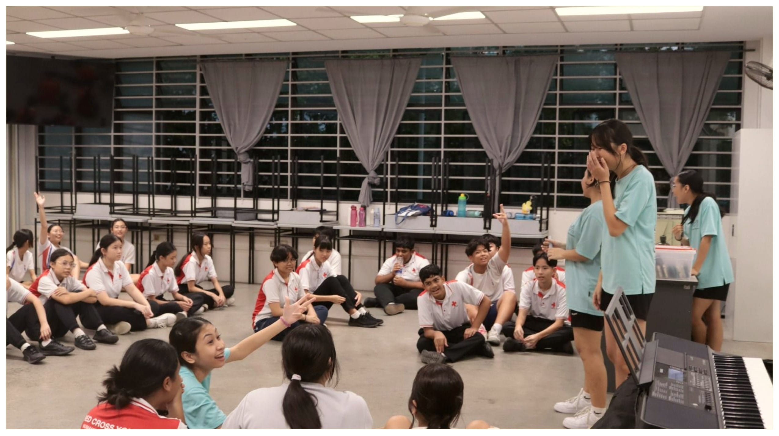 Students in white uniforms sit on a floor in a circle while two instructors in teal shirts lead an activity near a keyboard.