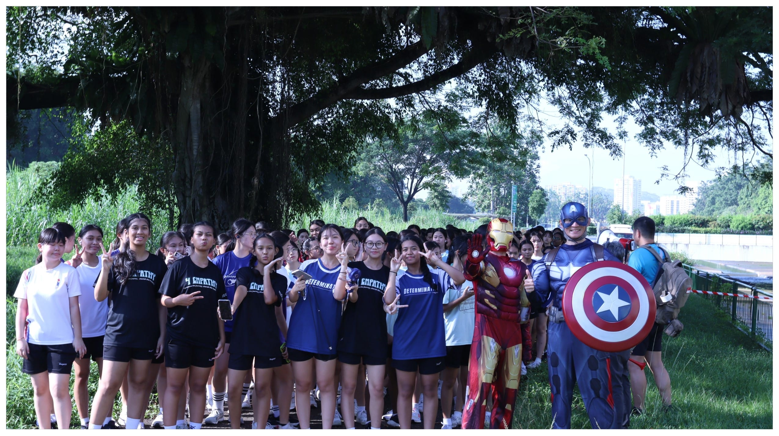 A large group of students in blue shirts pose outdoors with people in Iron Man and Captain America costumes under a large tree.