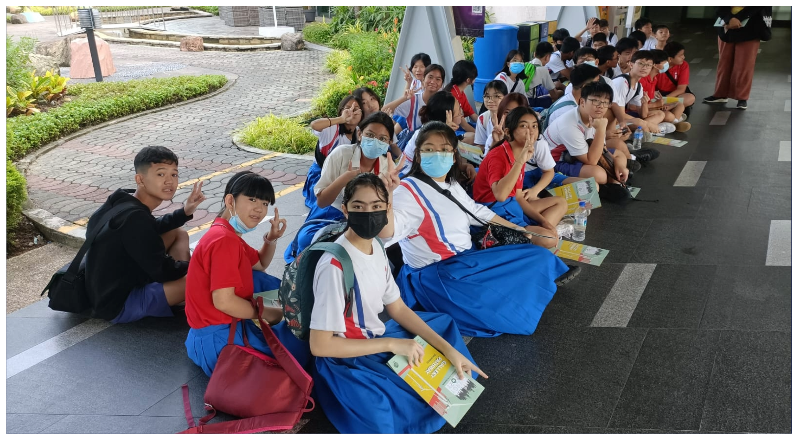 Students in uniforms sit in a long line on the floor, some wearing masks and flashing peace signs.