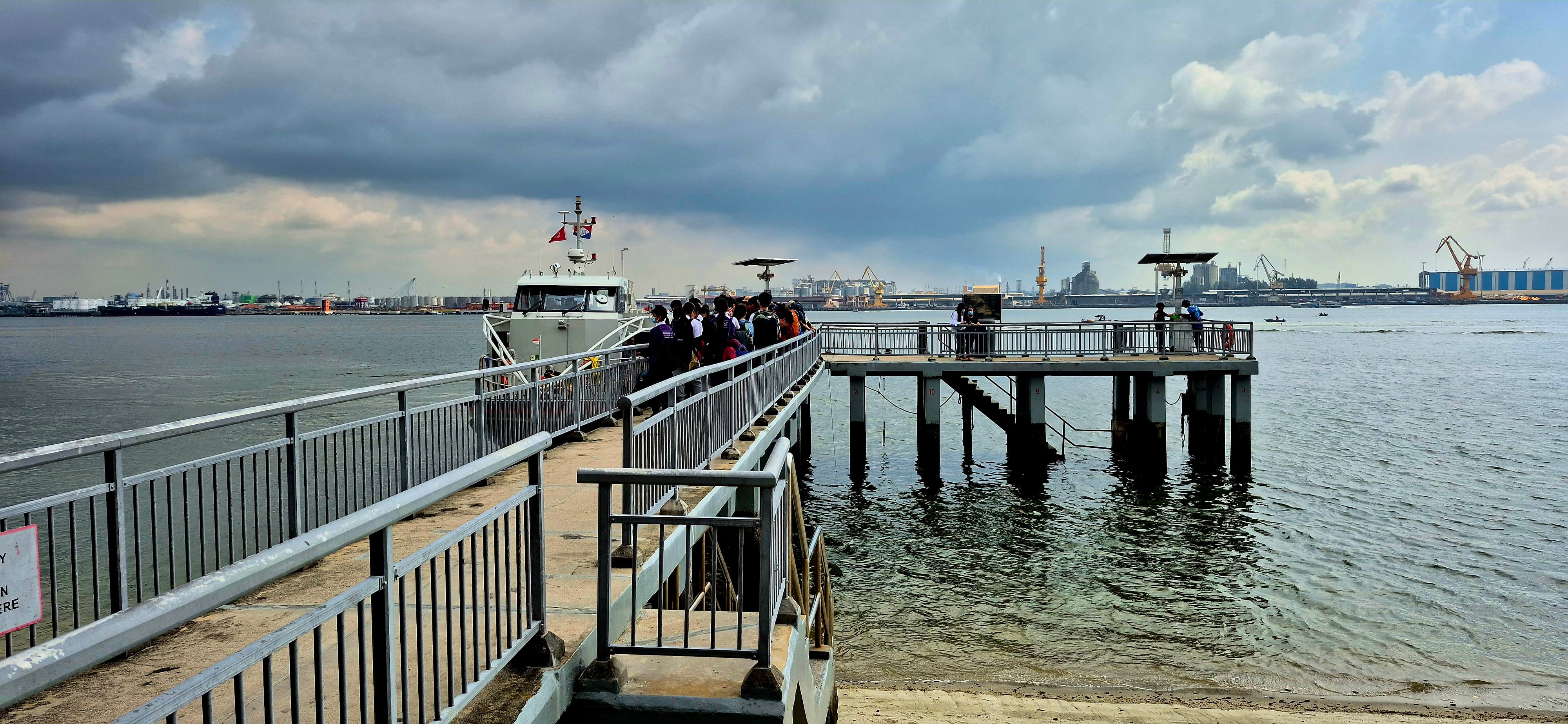 People boarding a vessel at a waterfront pier under dark clouds, with an industrial port skyline in the background.