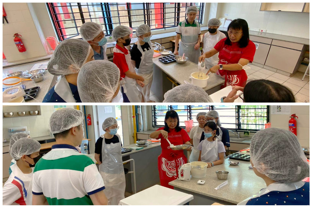 A woman in a red apron demonstrates baking to a group wearing hairnets and aprons in a kitchen classroom.