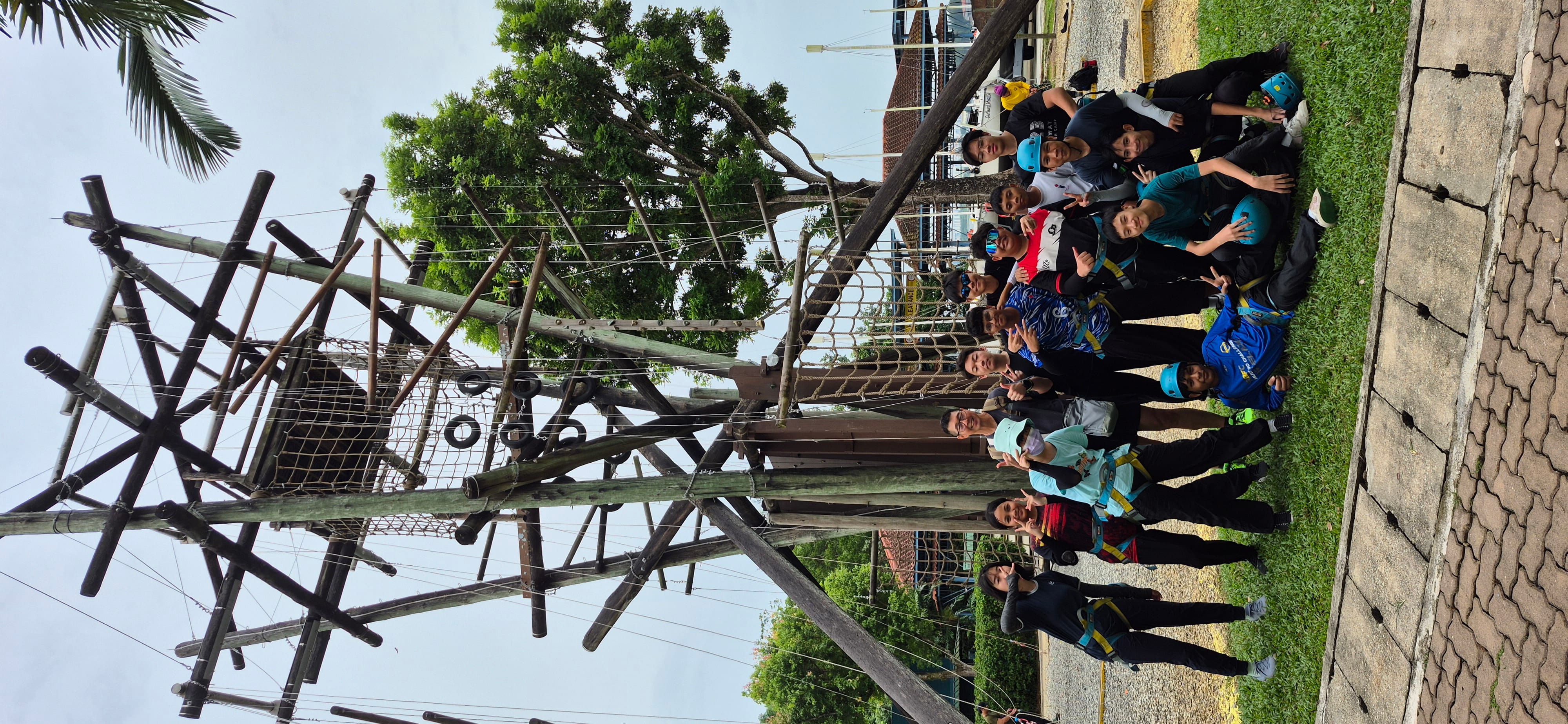 A group of teens in harnesses and helmets pose together in front of a tall wooden rope climbing tower outdoors.