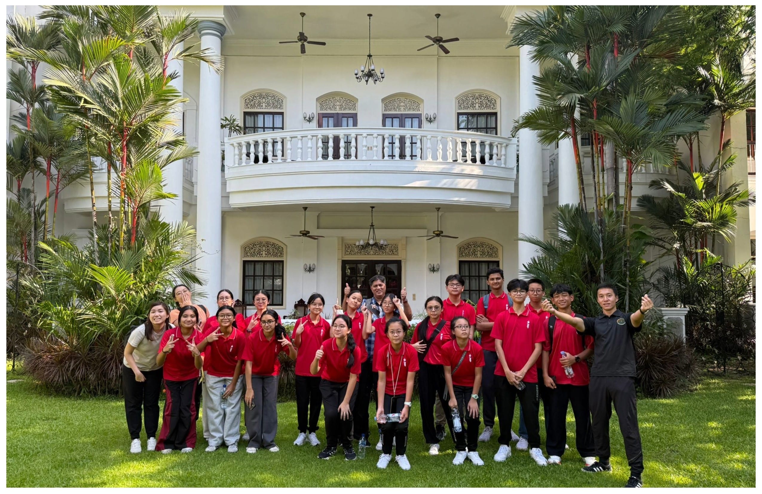 A group of about 20 students in red polo shirts pose on a lawn in front of a white colonial-style building with a balcony.