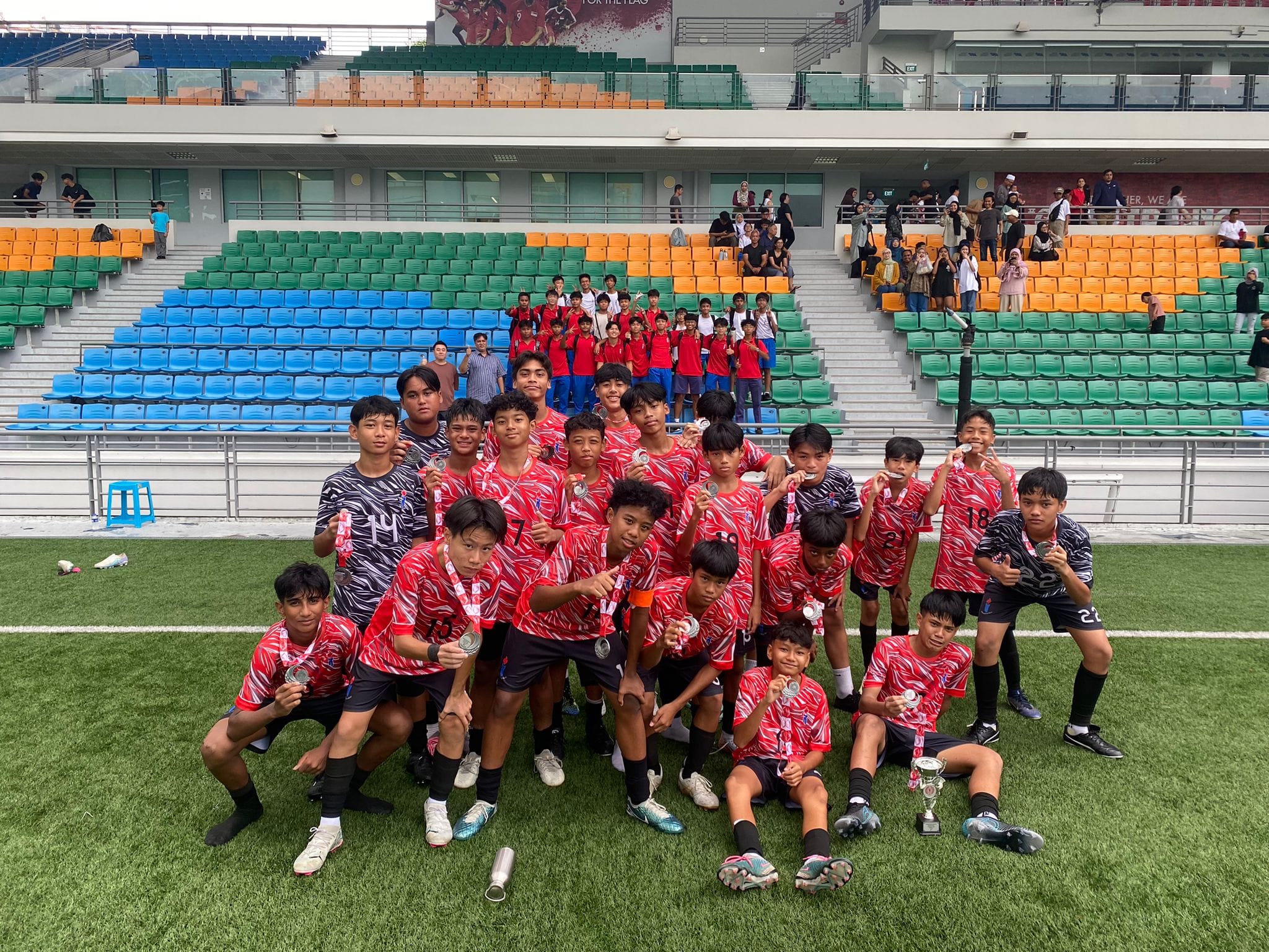 A youth football team in red jerseys poses on a stadium pitch, holding medals and a small trophy.