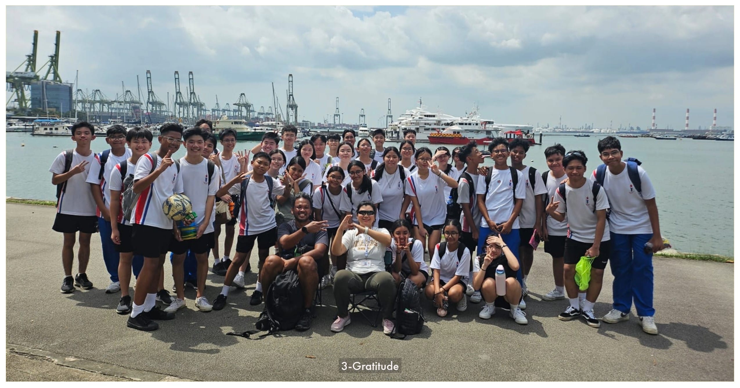 Large group of students in white uniforms posing outdoors by a harbor with cranes and boats in the background.