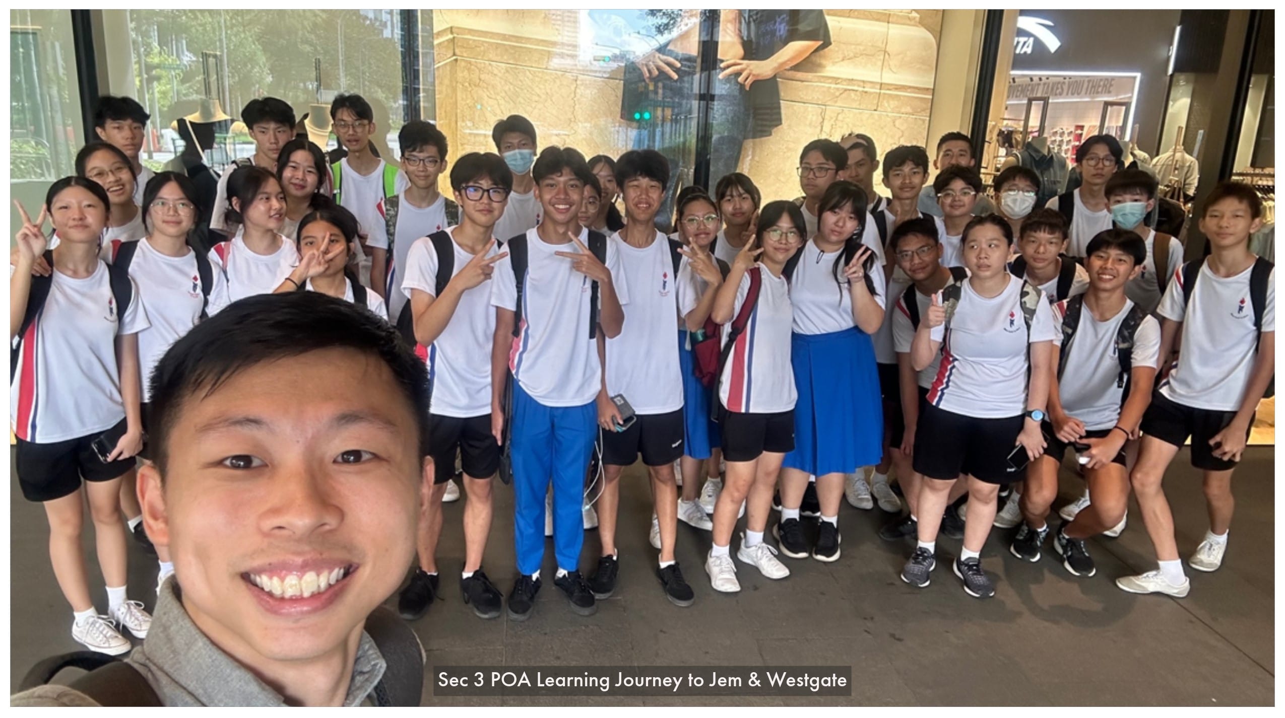 A teacher takes a selfie with a large group of students in white school uniforms posing outside a mall.